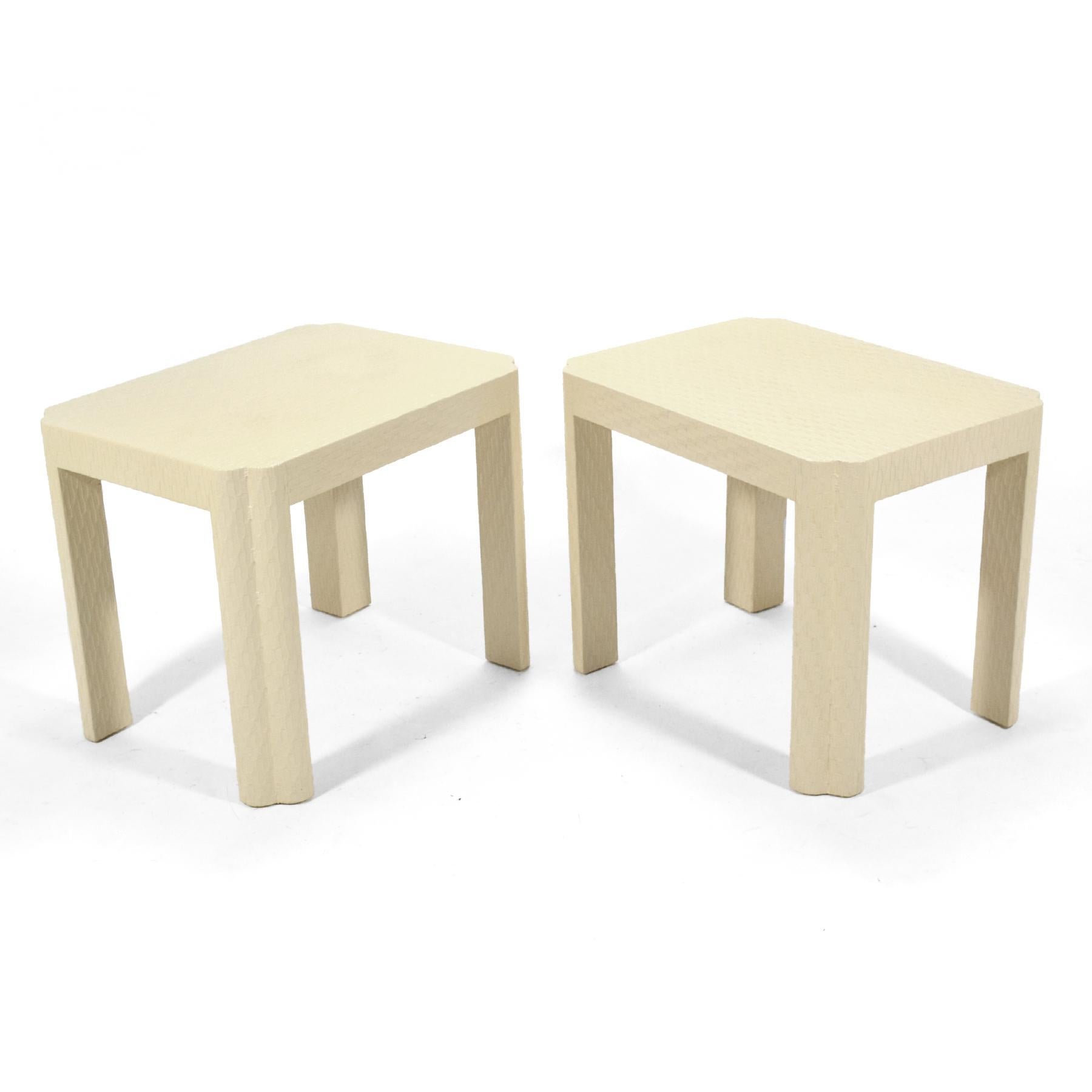 This pair of handsome side tables are very similar to the designs of Karl Springer. They are wrapped in a textured fabric, painted a warm ivory color, and have sculpted corners/ legs. Possibly made by Baker Furniture Co. they are unmarked.