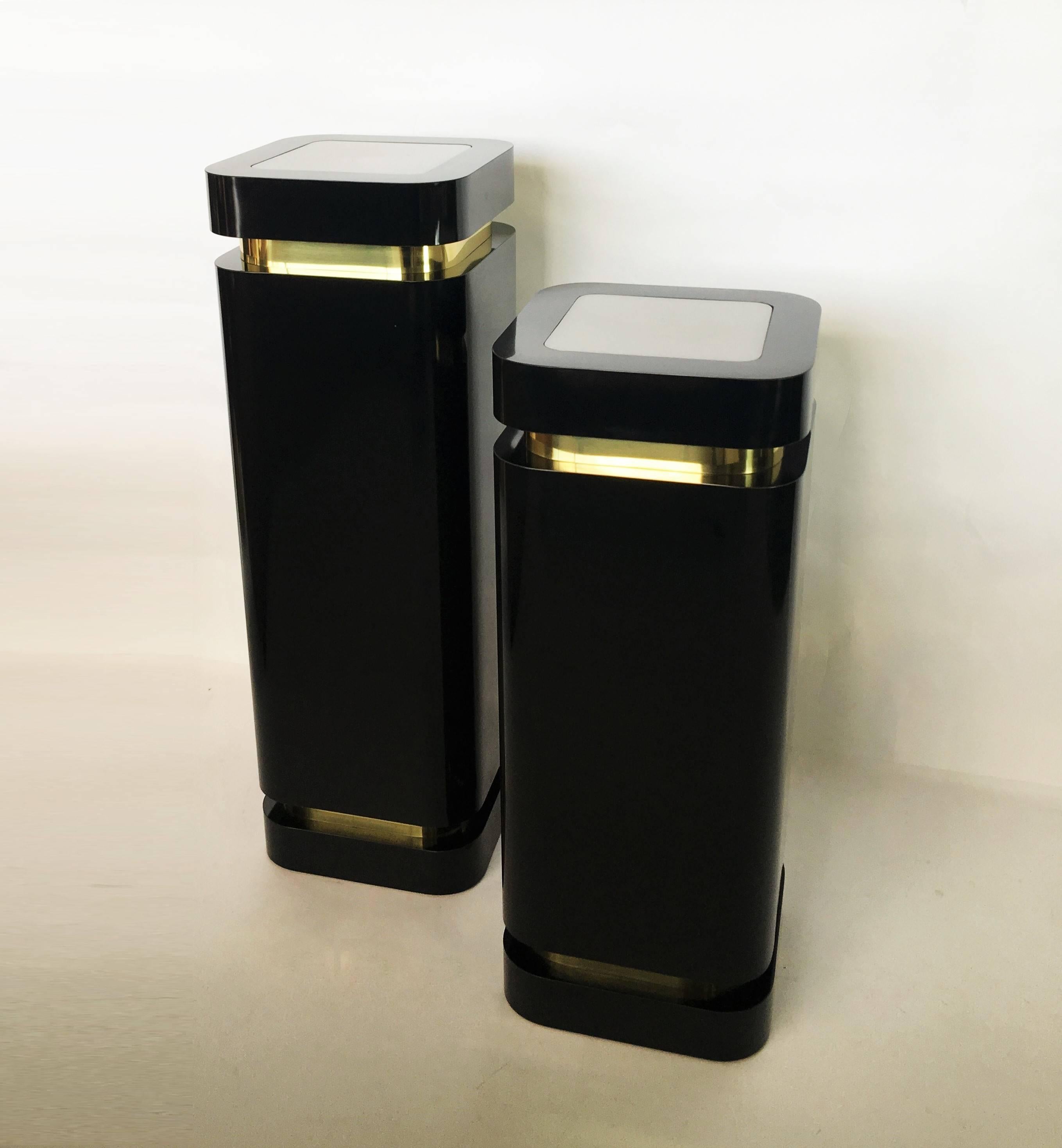 Reminiscent of works by Karl Springer with the signature curved edges and Art Deco inspiration. Two unique black laminate lighted display pedestals with brass trim. Tops with up lights are acrylic plexi-glass in frosted white. One is slightly higher