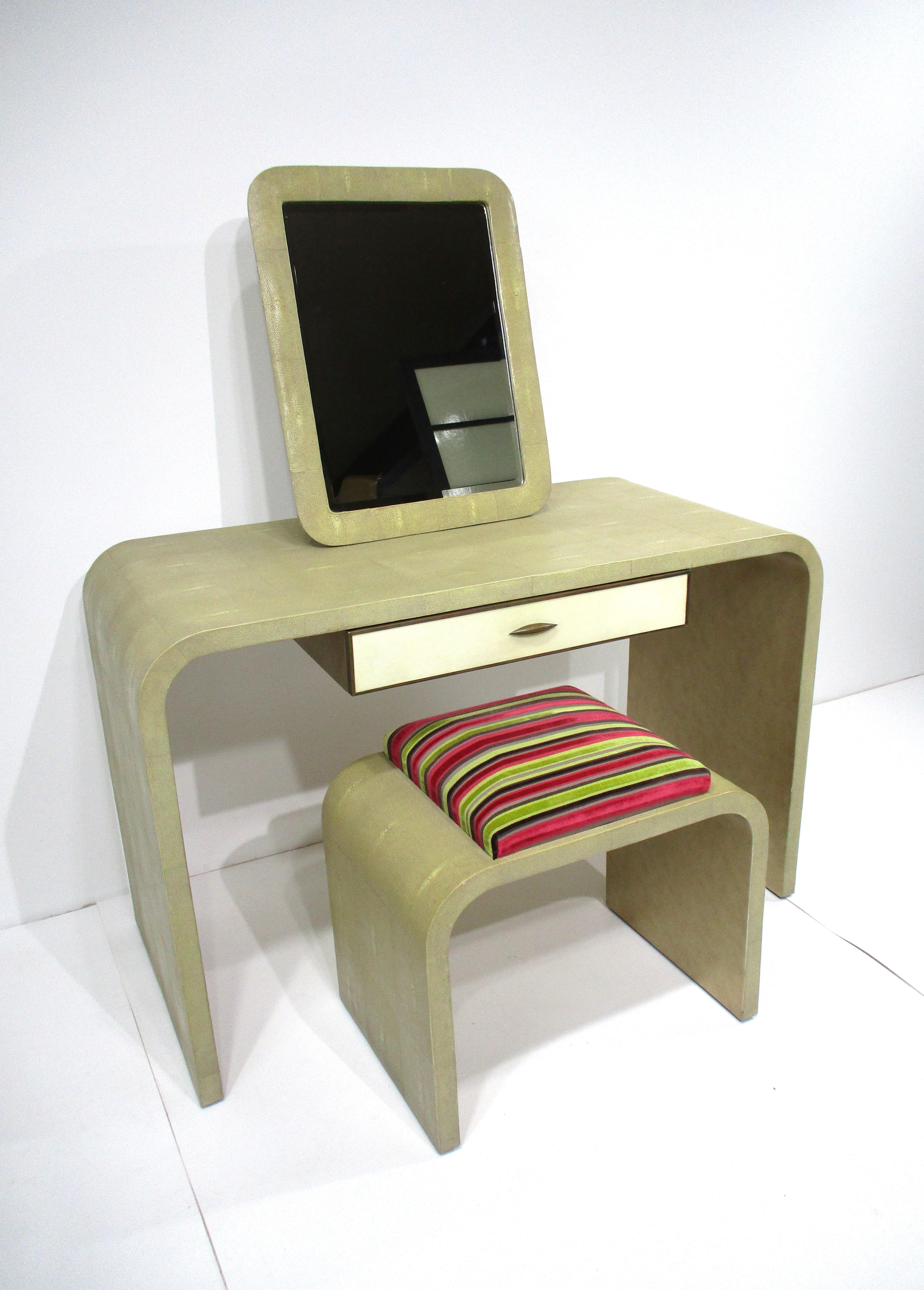 A luxurious three piece shagreen covered vanity set with matching stool having Jacobs coat fabric on the upholstered seat . A free standing mirror tops the vanity table which also has a drawer with the handle and edges of it in a bronze metal . This