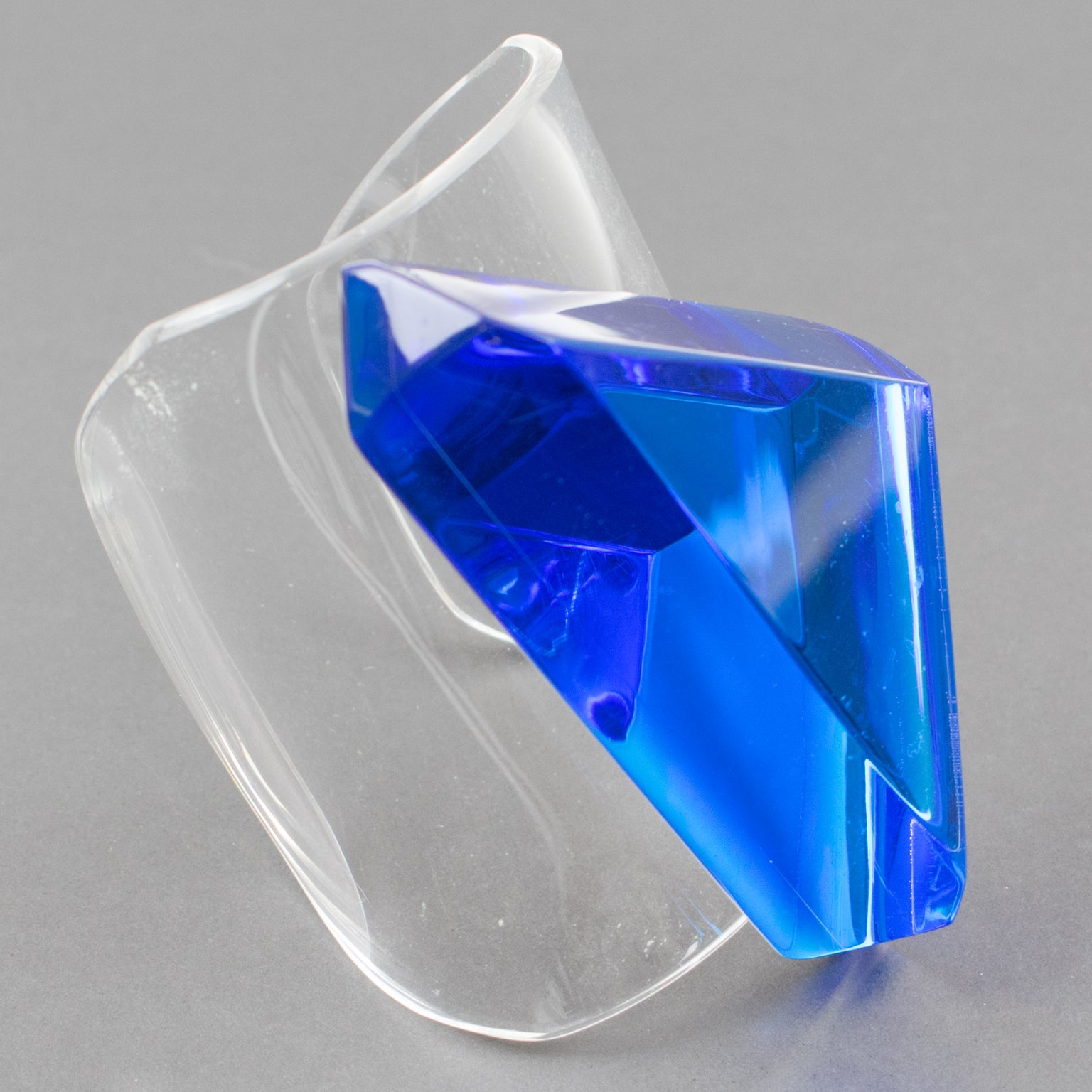 A striking Lucite cuff bracelet designed by Harriet Bauknight for Kaso. Clear Lucite oversized rigid cuff shape topped with large geometric ornament. The Lucite ornament features a dimensional geometric faceted large triangle in intense blue lagoon