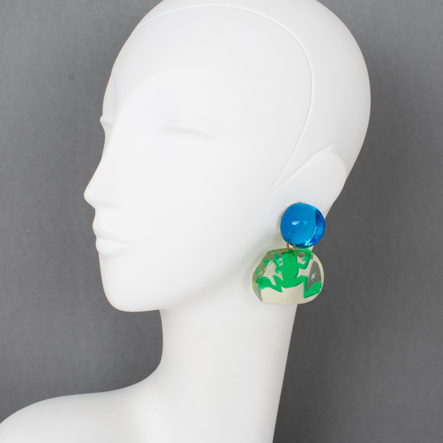 These are the cutest Lucite dangling clip-on earrings Harriet Bauknight designed for Kaso in the 1980s. They feature a lagoon blue Lucite dimensional bead fastening complemented by a rounded transparent pebble element in which a green frog is