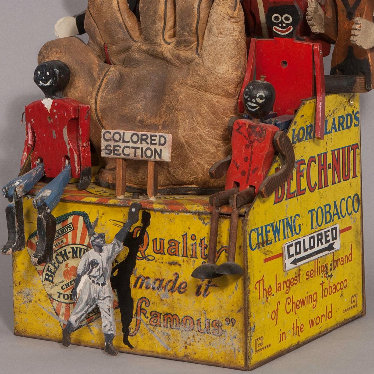 assemblage sculpture: chewing tobacco tin remade into baseball stands, 2 handmade scoreboards, old glove, carved baseball figure, old wood racist caricatures of people in the stands

This baseball imagery symbolizes a racist aspect of American