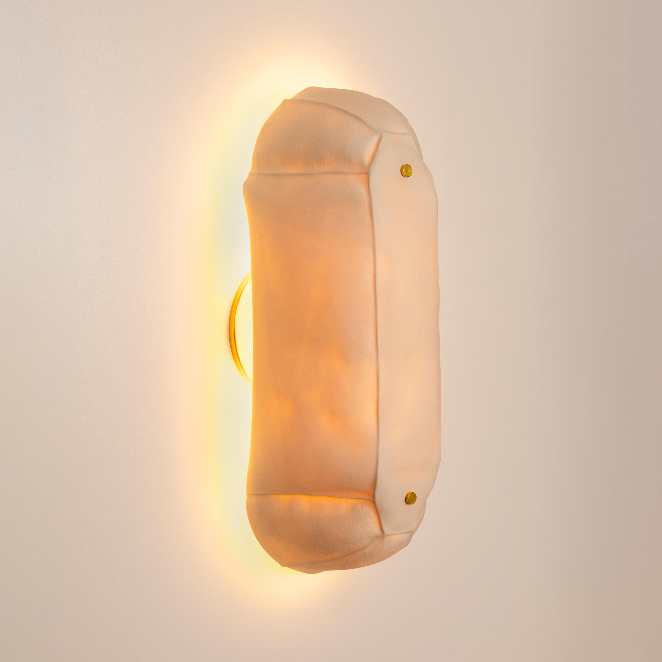 Tactile and textured porcelain wall sconce light in a bulbous racetrack shape. The translucent porcelain diffuser casts a warm-colored glow when lit. 

Shown in the photos with a waxed brass canopy and hardware. Custom metal finishes are available