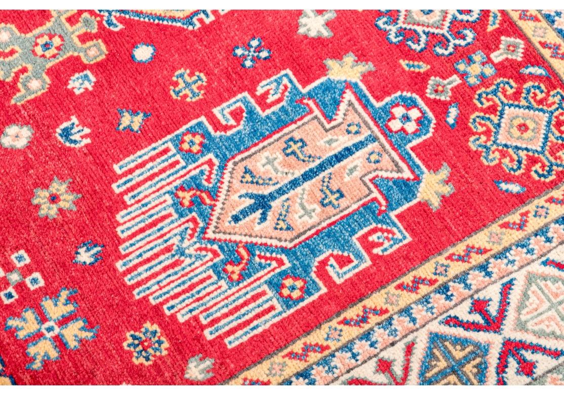 Handgeknüpfter Wollteppich mit einem zentralen blauen Rautenmotiv, umgeben von 4 gegenüberliegenden abstrakten Motiven mit mehrfarbigen Abstraktionen dazwischen. Hinzu kommen verstreute Blumen, abstrakte Motive und Sterne auf einem roten Feld. Der