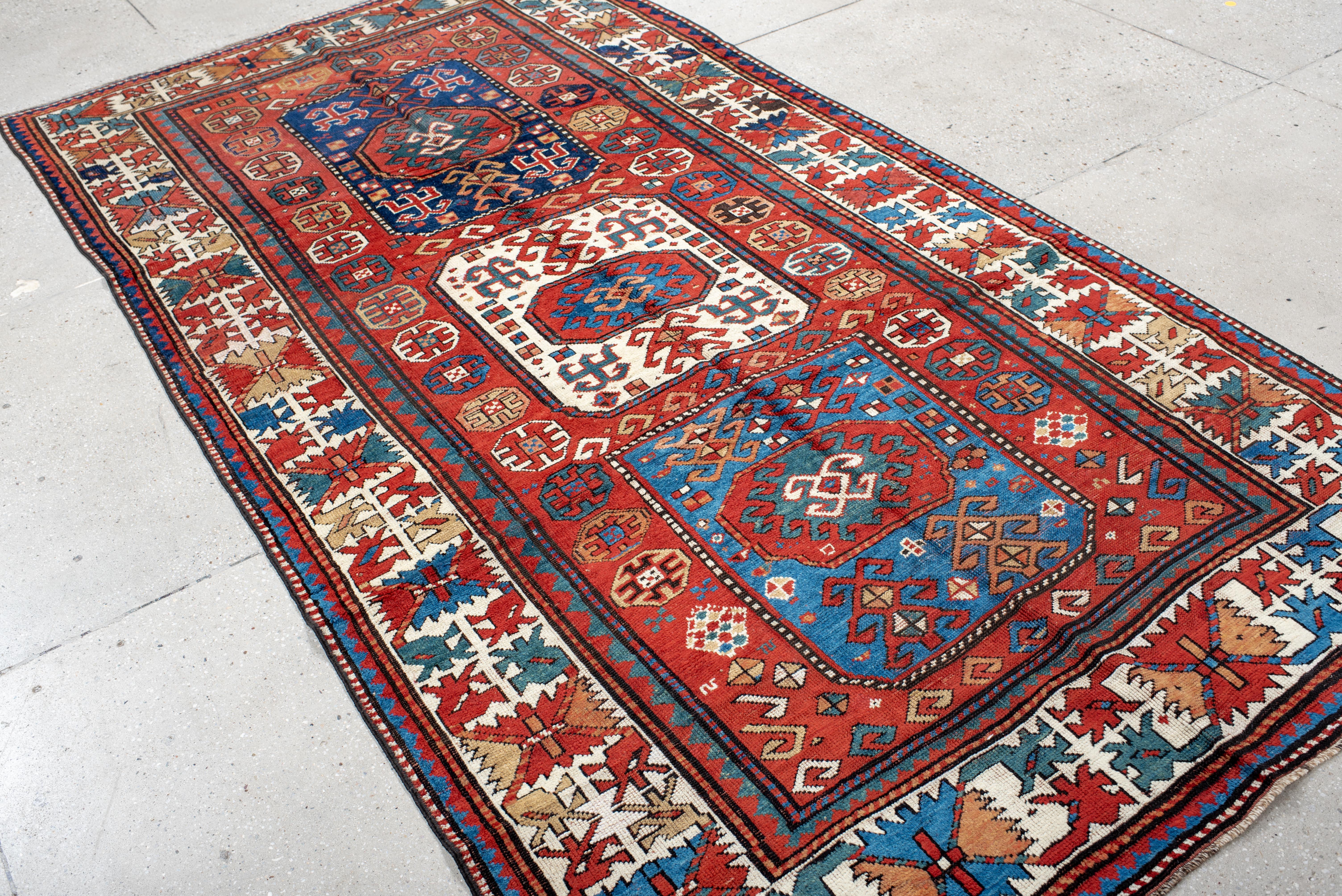 This antique Kazak rug exemplifies the bold geometry and rich coloration of the Caucasus weaving tradition, featuring a striking vertical arrangement of three medallions anchored within a vibrant red field. Indigo, ivory, and azure blues contrast
