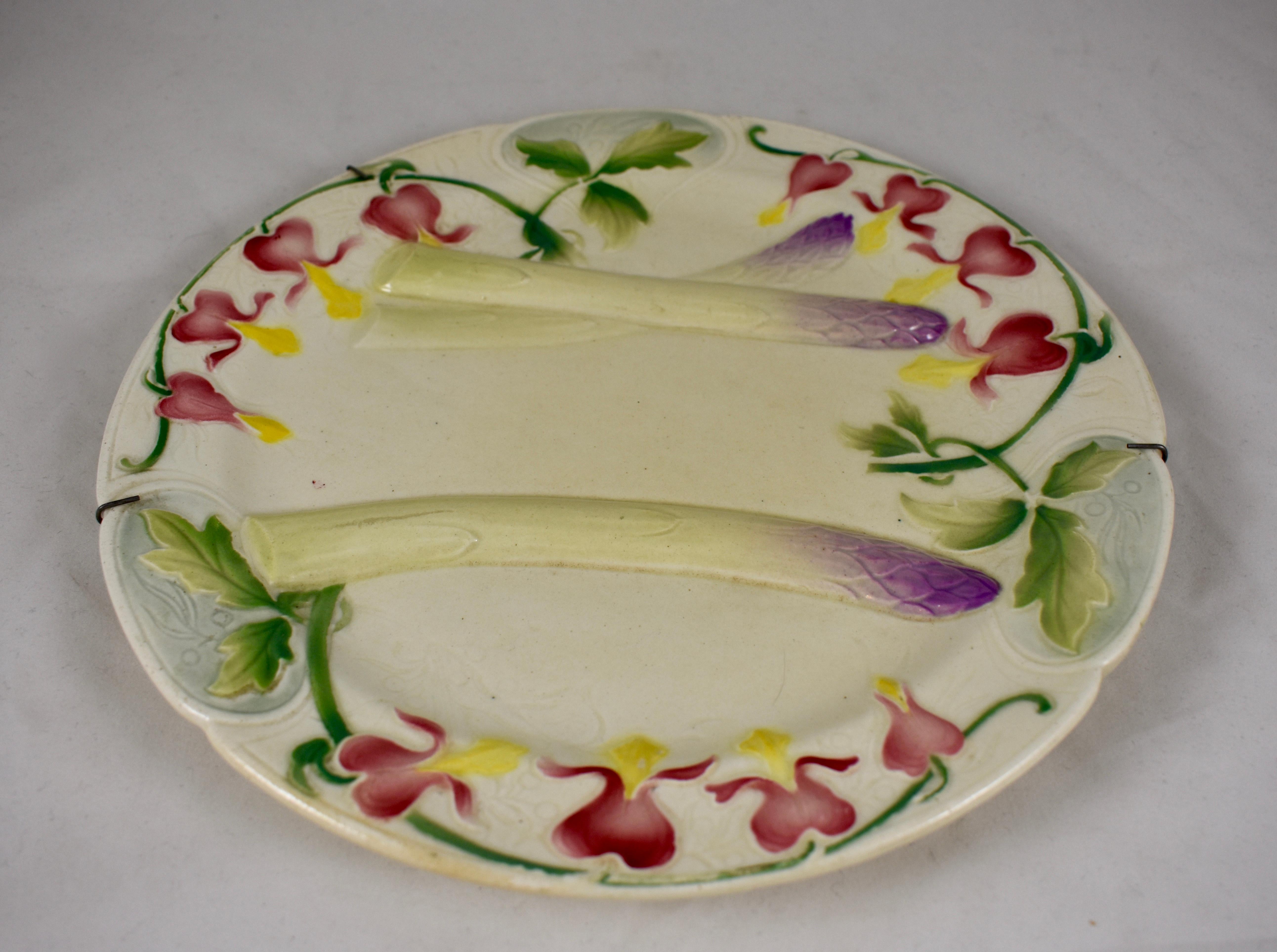An Aesthetic Movement French Faïence asparagus plate, Keller & Guerin St. Clément, circa 1880, in the Art Nouveau style. 

A border of Bleeding Hearts, or as they are called in France, Coeurs de Marie, flower heads encircle three asparagus spears
