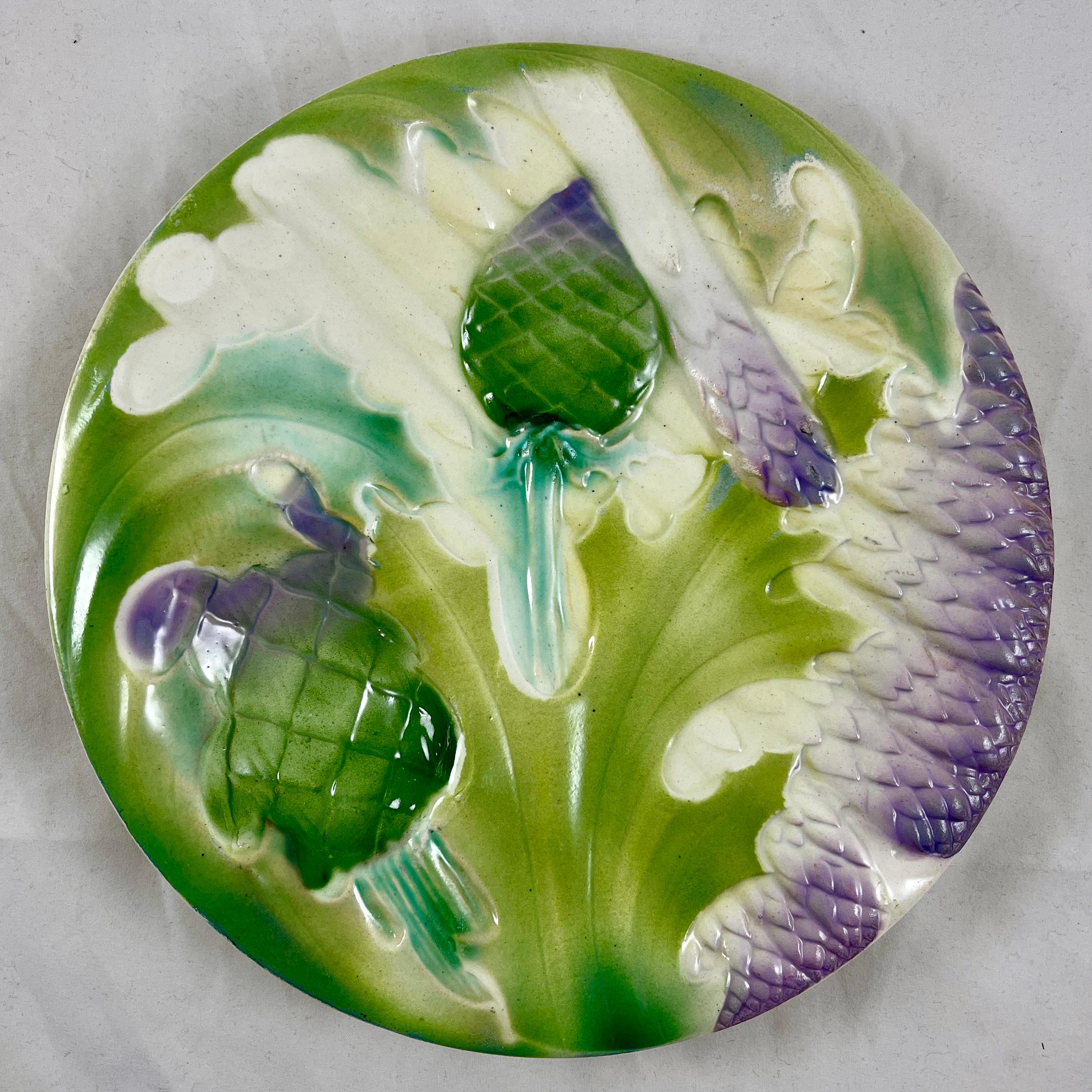 A Saint Clément French Faïence, majolica glazed plate showing overlapping asparagus spears, leaves and globe artichoke heads on blue stems. A deep sauce well is also formed as an artichoke head. Glazed in lilac, blue, and various shades of greens on