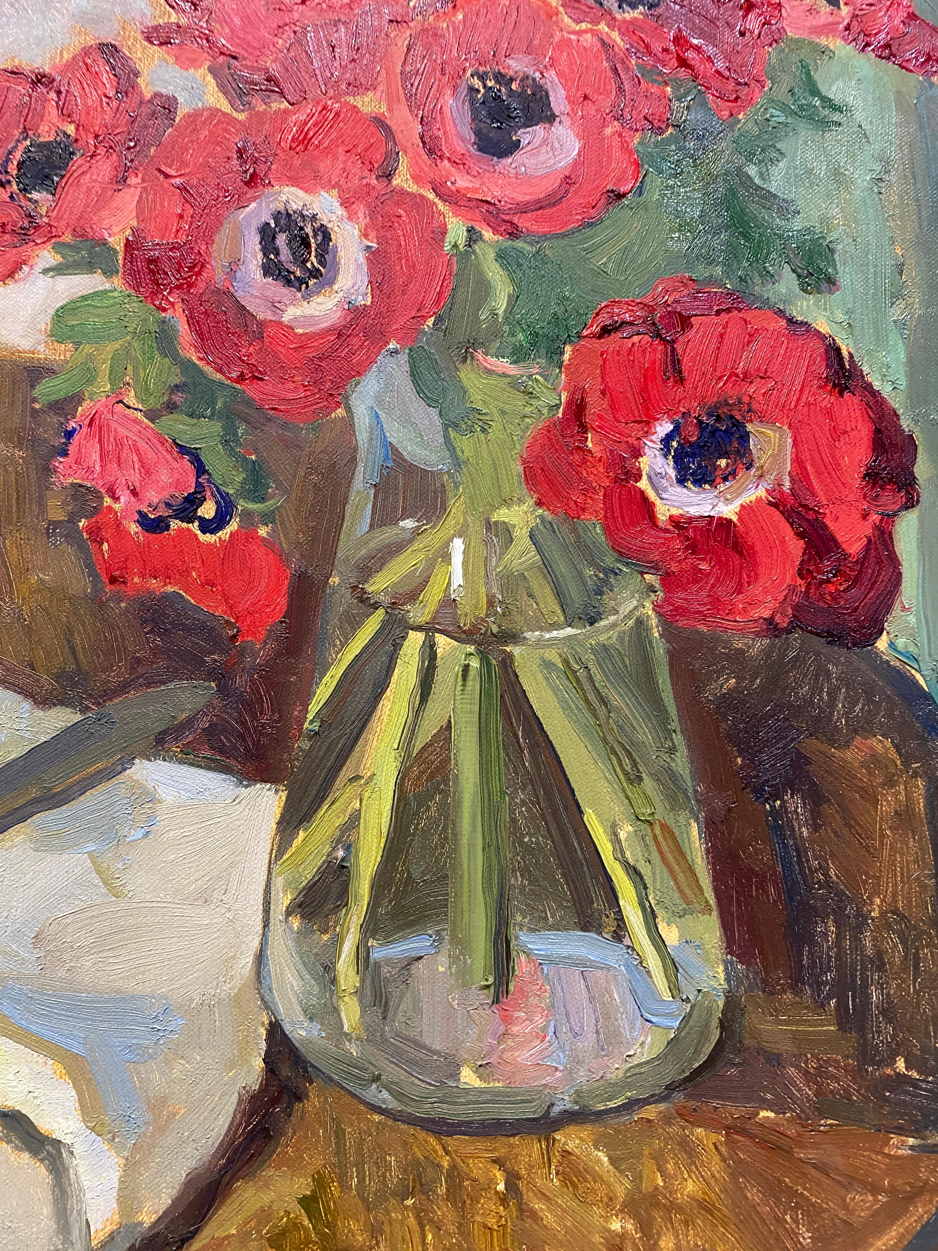 A still life painting of red anemones in a glass vessel, situated on a small round wooden table. A butter knife rests upon neutral toned fabric. A green drape with red fringe trim hangs in the background. 

Framed in a white wood frame.

Framed