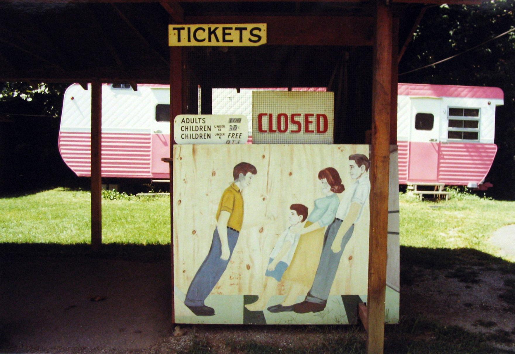 Pink Trailer Tilt, North Carolina by Ken Brown is a 9 1/4 x 13 1/2 inch Ektacolor print. This photograph features a ticket booth with painted leaning figures, in front of a pink and white trailer. This photograph is titled and signed with the