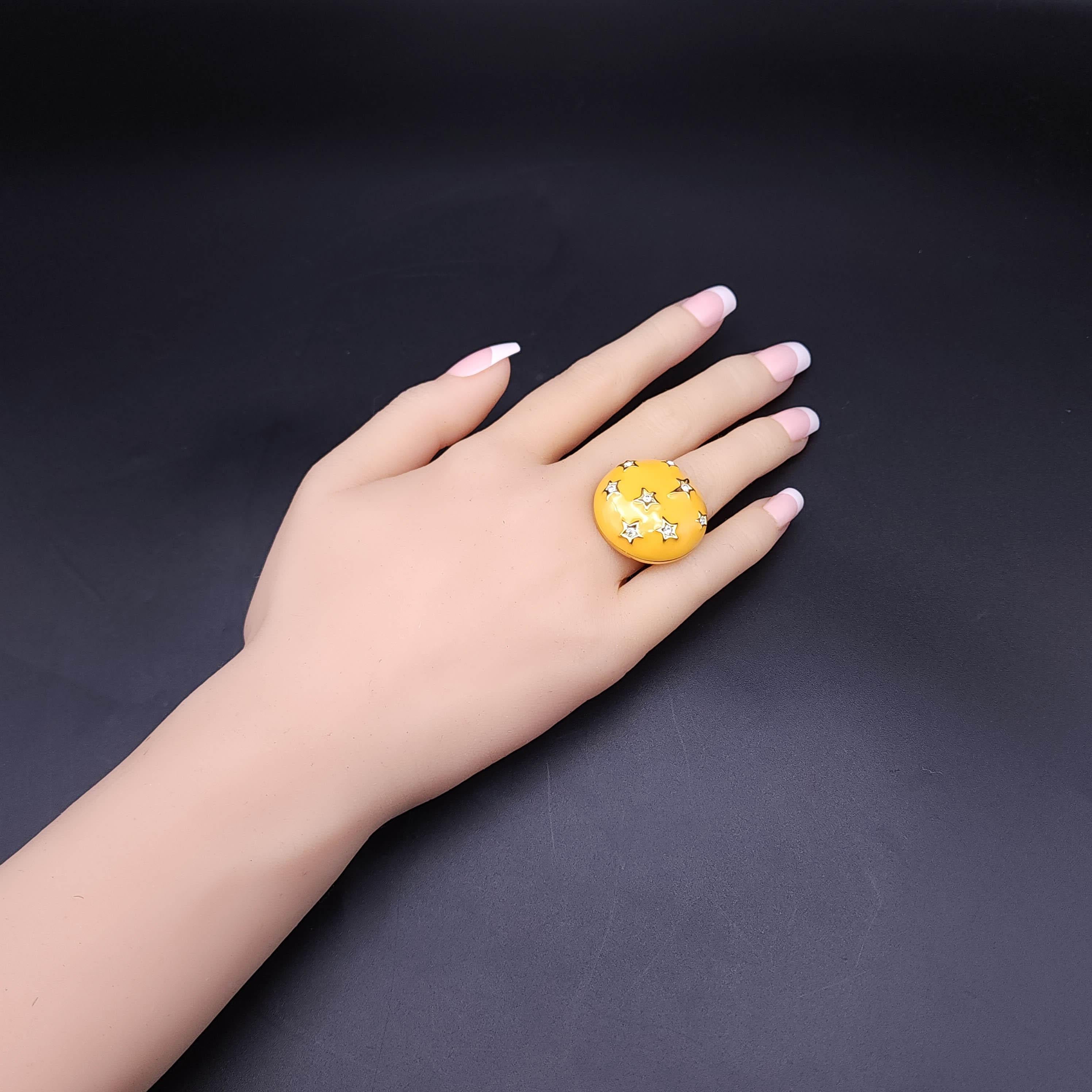Añade un toque de sol a tu conjunto con este anillo de cóctel de Kenneth Jay Lane. Esta vibrante pieza luce un radiante engaste de esmalte amarillo, adornado con estrellas doradas que acunan sendos cristales transparentes. La banda ajustable