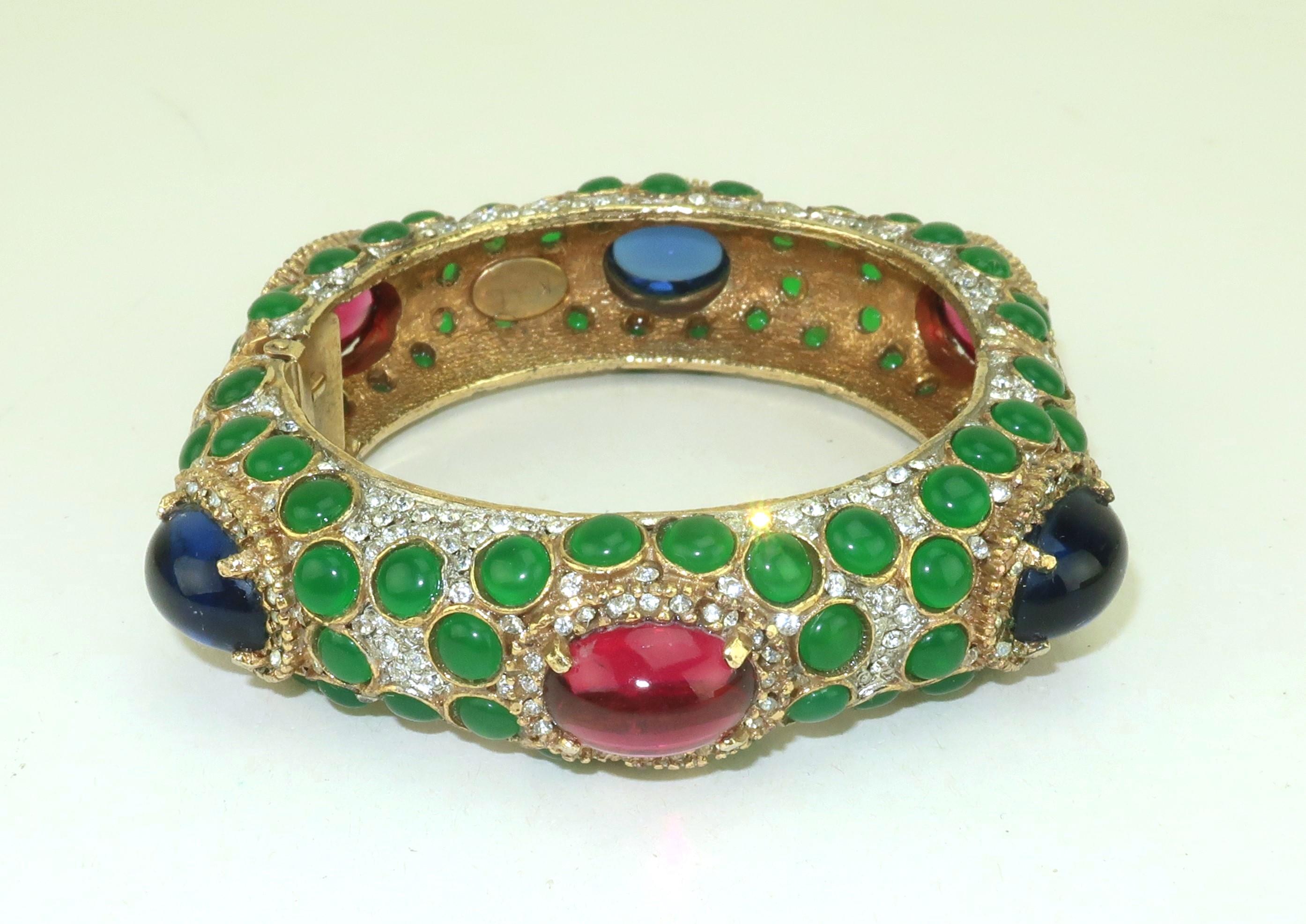 Bracciale bangle anni '60 di Kenneth Jay Lane in tonalità oro splendidamente impreziosito da pietre di vetro cabochon dall'aspetto di rubini, zaffiri e smeraldi cosparsi di strass di cristallo che imitano i diamanti.  Lo stile opulento ricorda gli