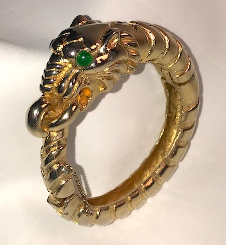 Early Kenneth Jay Lane tiger bangle in shiny gold plate. The head is set with green glass cabochon eyes. The mouth is open holding a ring that the tail of the tiger goes through and curves back. Hinge on one side and push pin closure on the opposite