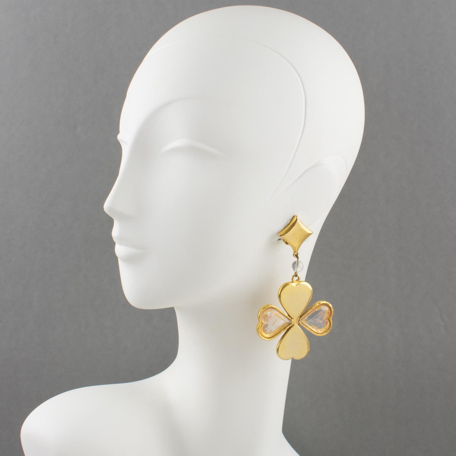 These lovely Kenzo Paris signed clip-on earrings feature a long dangling drop shape with a four-leaf clover design and heart-shaped petals. The gilt metal framing is all textured with a beveled glass insert. The glass inserts have gold flakes