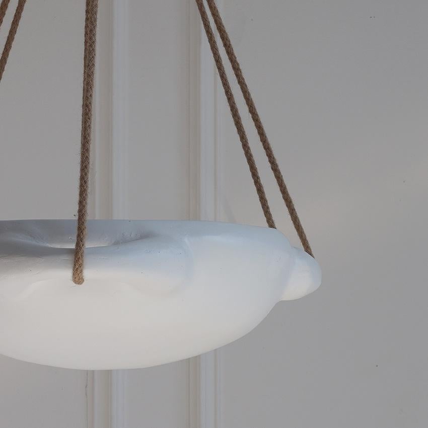 A gracefully suspended pendant, with a bowl-shaped shade crafted from fiberglass, and finished in smooth white plaster with a sculptural and organic aesthetic. Three braided natural rope cords extend from the top canopy to support the bowl, adding a