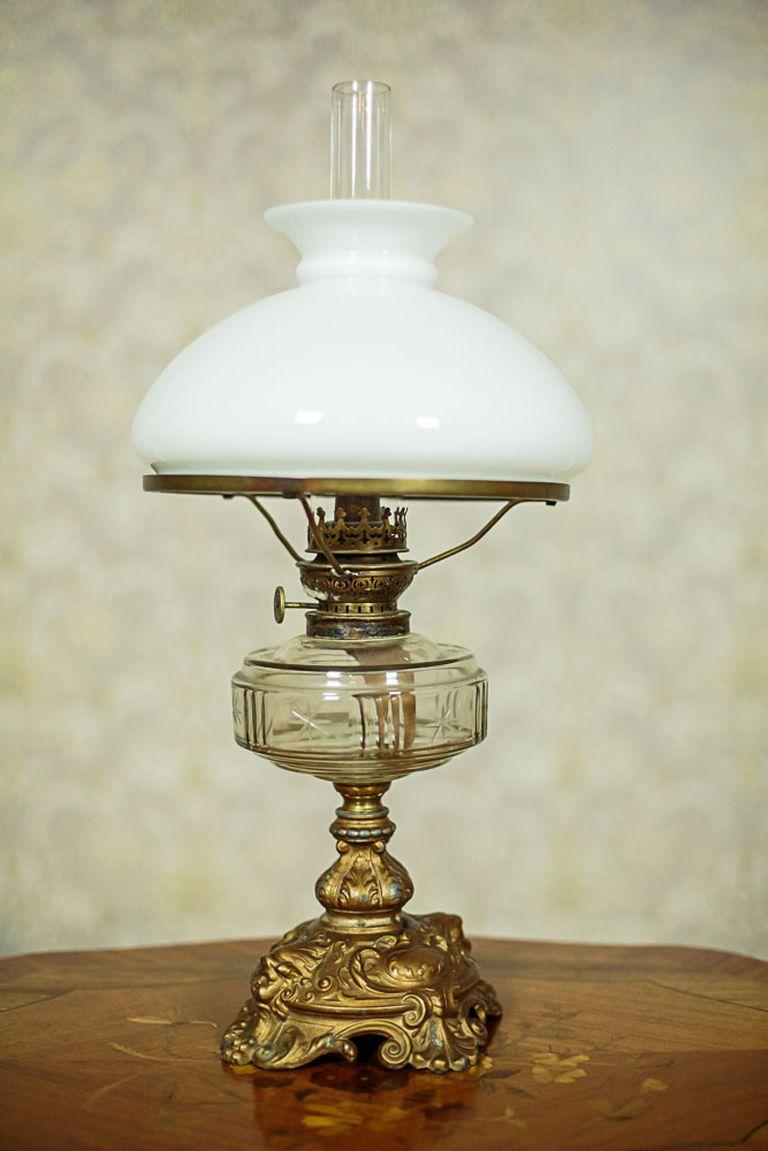 A lamp on a tin, cast foot that is covered with bronze.
The kerosene container is glass, with an engraved pattern.
The lampshade is made of milky glass.

Presented lamp is in good condition. There are traces after soldering at the joining of the