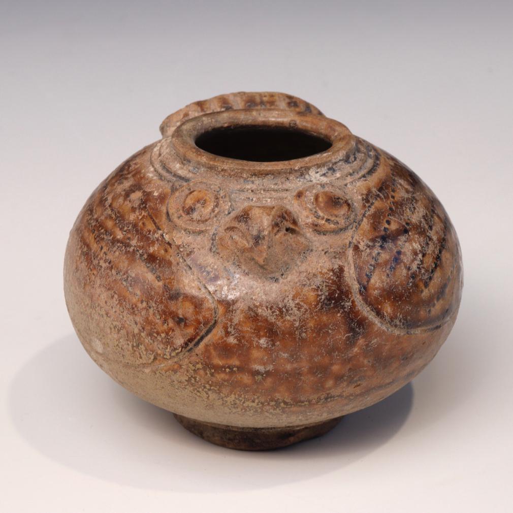 Khmer Ceramic Lime Pot. A globular stoneware jarlet in the form of a bird covered in a caramel color glaze stopping short of the elevated and slightly convex string  cut foot displaying the beige biscuit underneath. A owl’s beak and protruding eyes