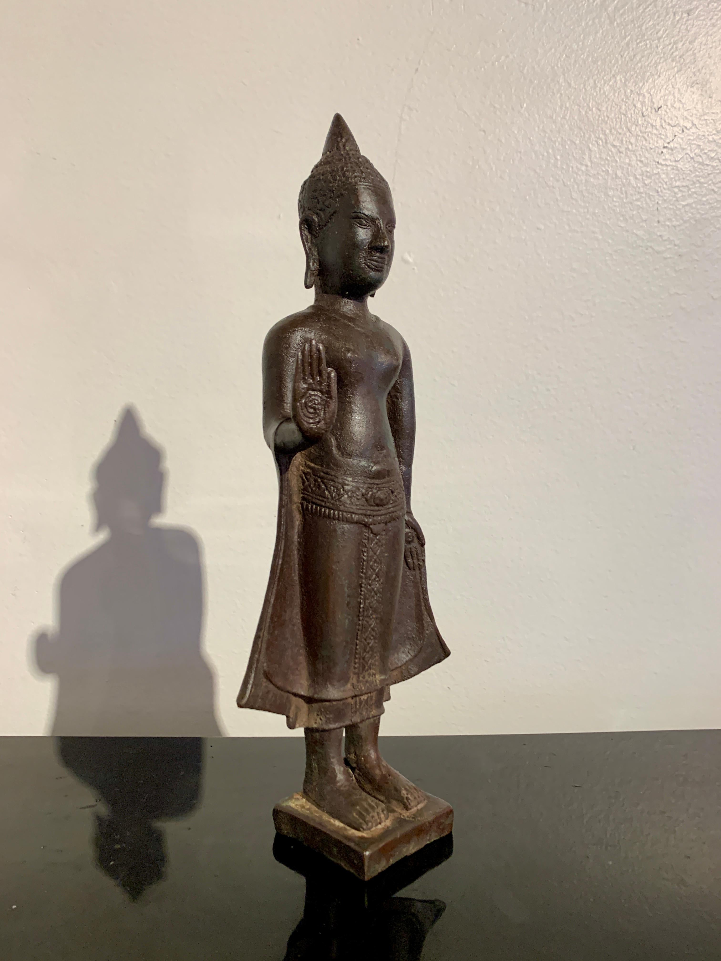 Une belle figure de style khmer vintage en bronze moulé et patiné d'un Bouddha debout, milieu du 20e siècle, Thaïlande ou Cambodge. 

Le Bouddha est représenté debout sur un socle carré, bas et uni. Il est vêtu d'un dhoti avec des ourlets décorés,