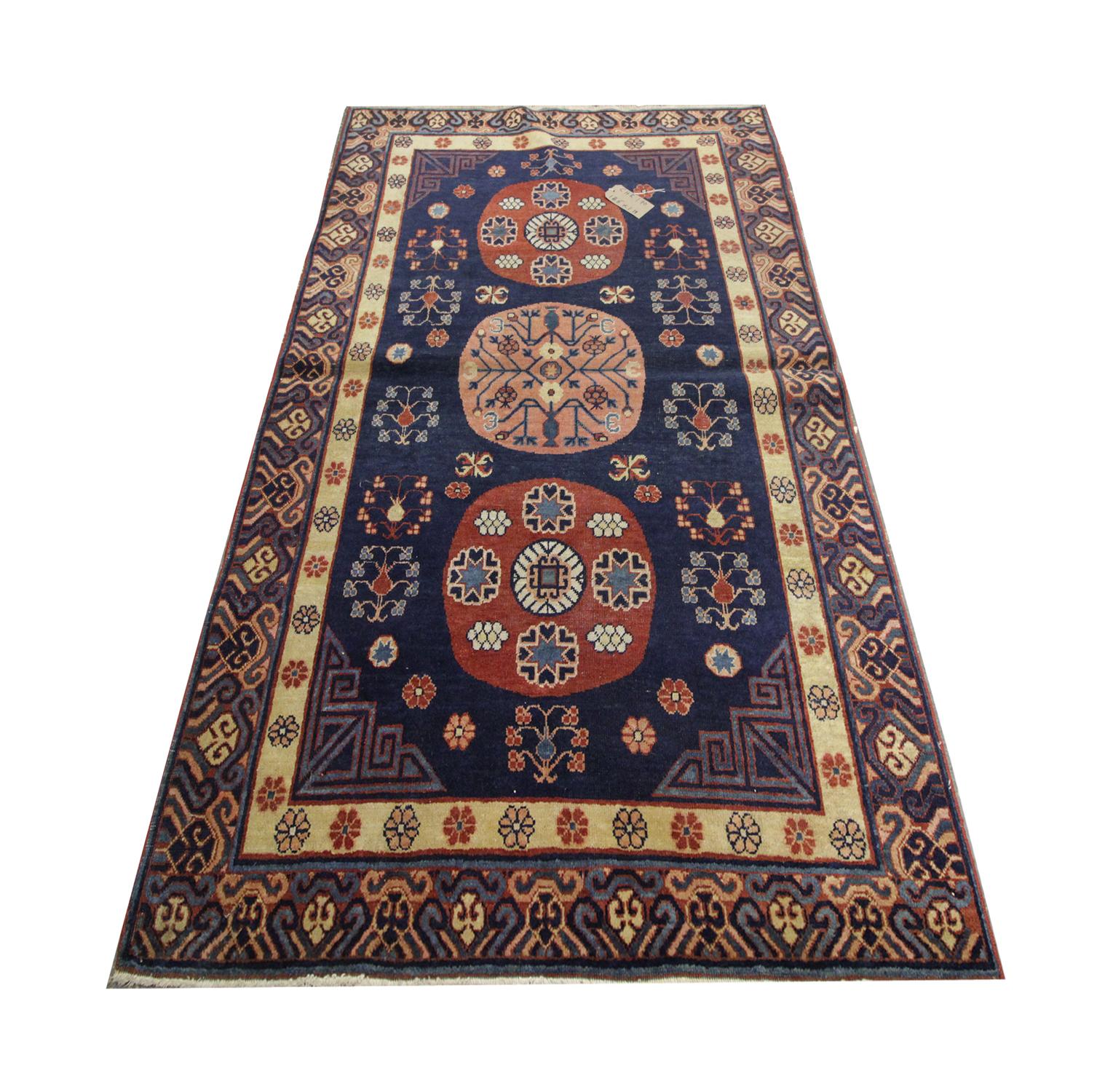 Ce tapis d'Orient bleu foncé d'Asie centrale a été tissé à la main à Kohtan en 1989. Trois emblèmes circulaires traversent le centre en rouge et pêche, entourés de motifs floraux géométriques. Il est entouré de motifs d'angle et d'une bordure jaune