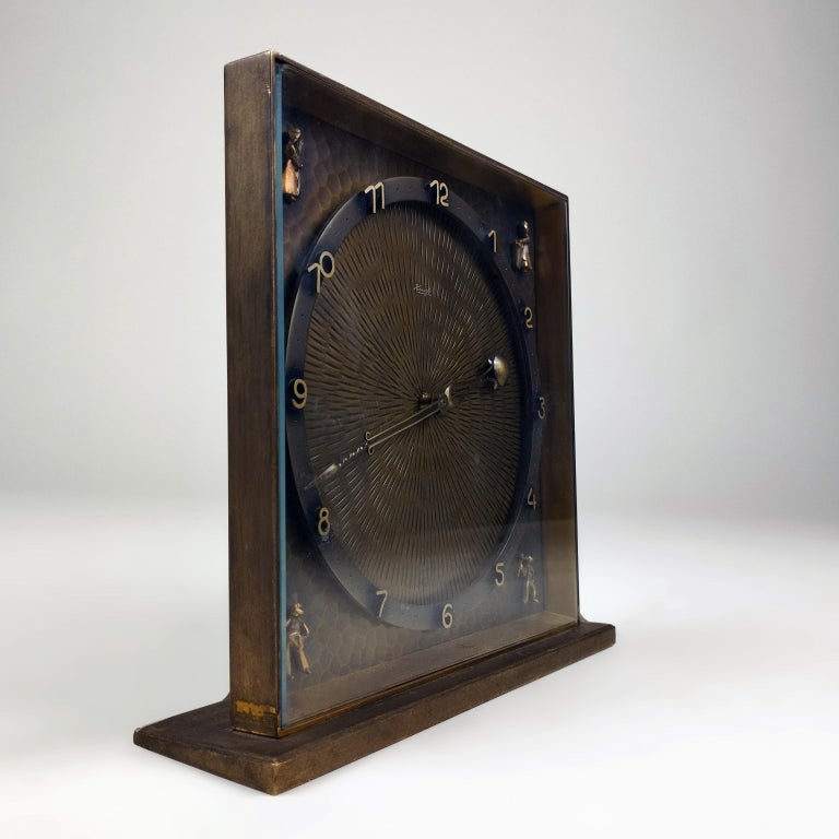 A rare German ca. 1960 bronze table clock by Kienzle. 

The squared 20.3 x 20.3 cm. box with front glass shows an inner 17 cm. Ø circle with the clock's dials. The 4 corners depict small peasant figures on a hammered background. The inner circle