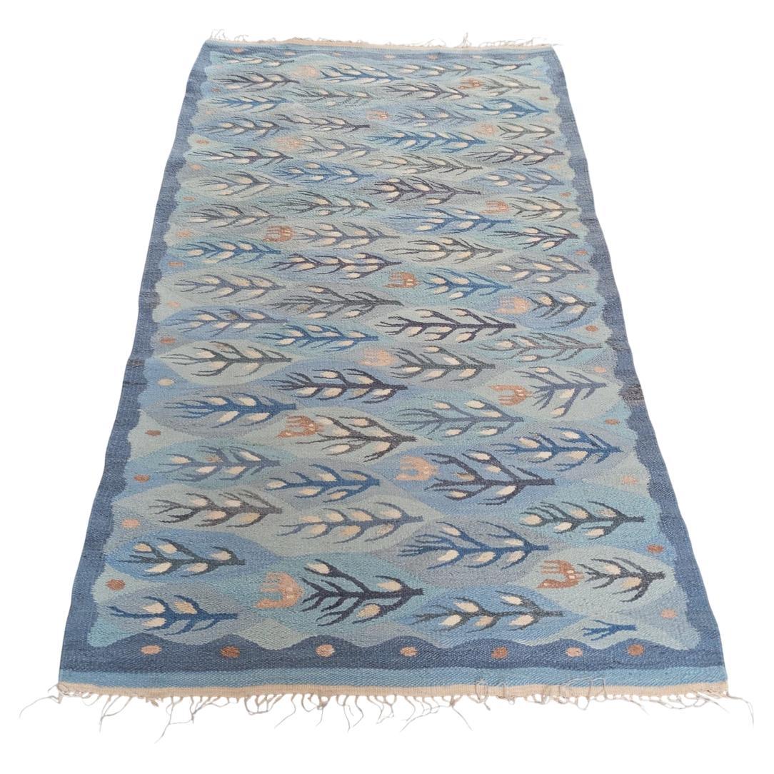 This rectangular-shaped carpet has a predominance of light blue and blue hues. The decorative motif consists of a series of stylized elements arranged in rows. Dark blue and blue trees with snow and some brown birds. The border has a blue frame with