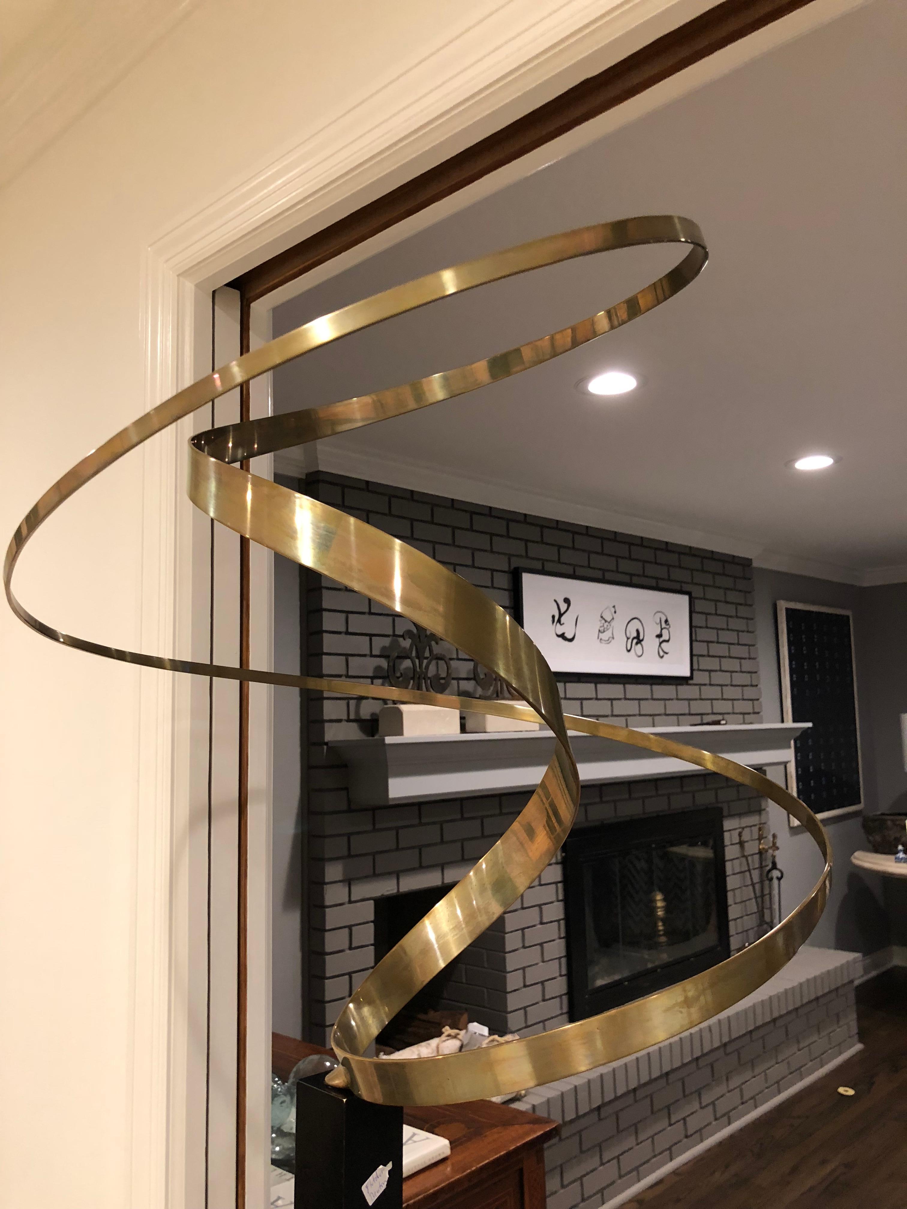 An abstract large brass ribbon sculpture  having Kinetic capabilities when plugged in. (It does not have to be plugged in to appreciate its presence.) Housed in a black metal stand. Cord is at base.