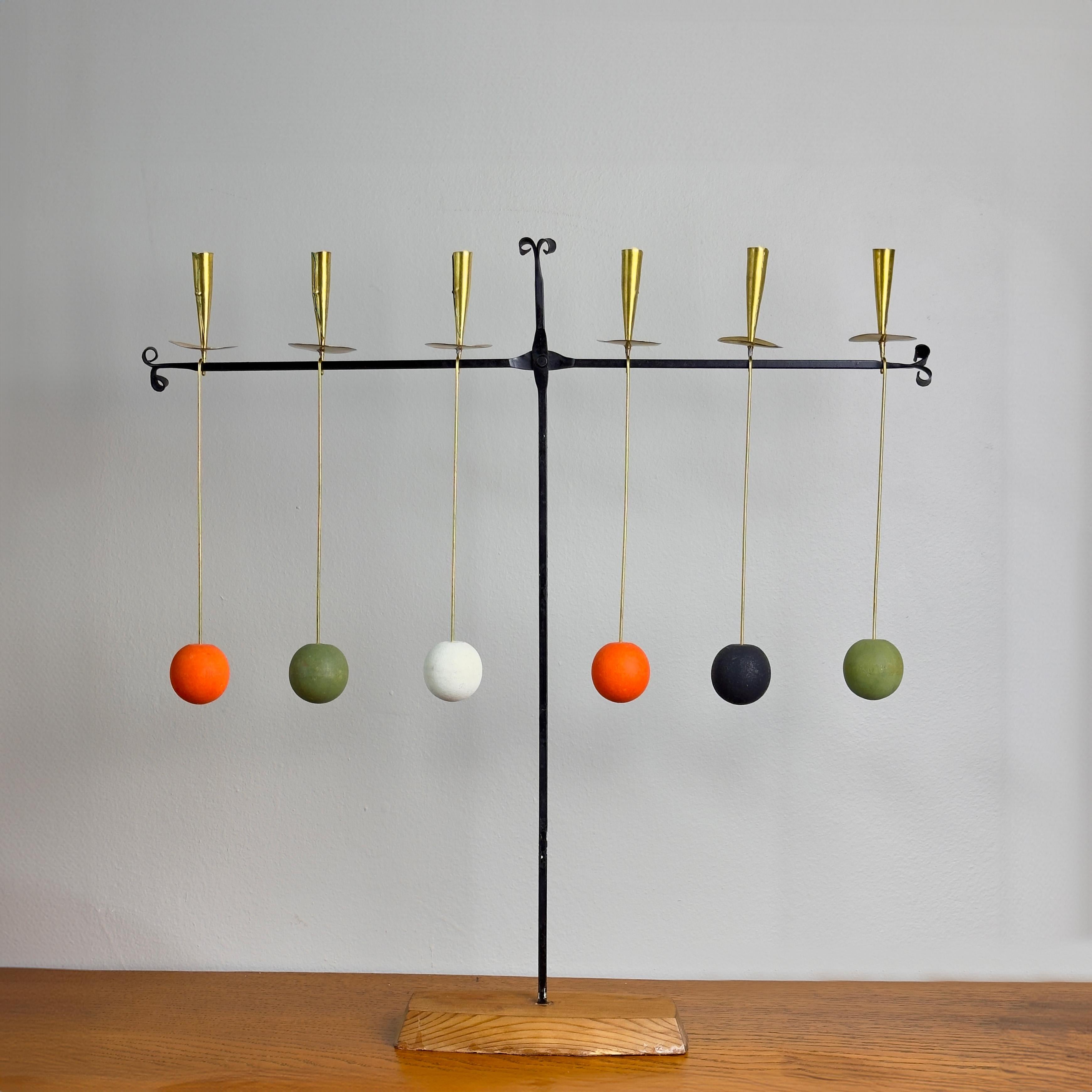 A handmade, kinetic candelabrum adorned with hanging brass candle holders counterweighted by painted wooden balls. 

This candelabrum is attributed to Swedish ceramicist, Thomas Hellström. The swirling wrought-iron tendrils and bright colours are