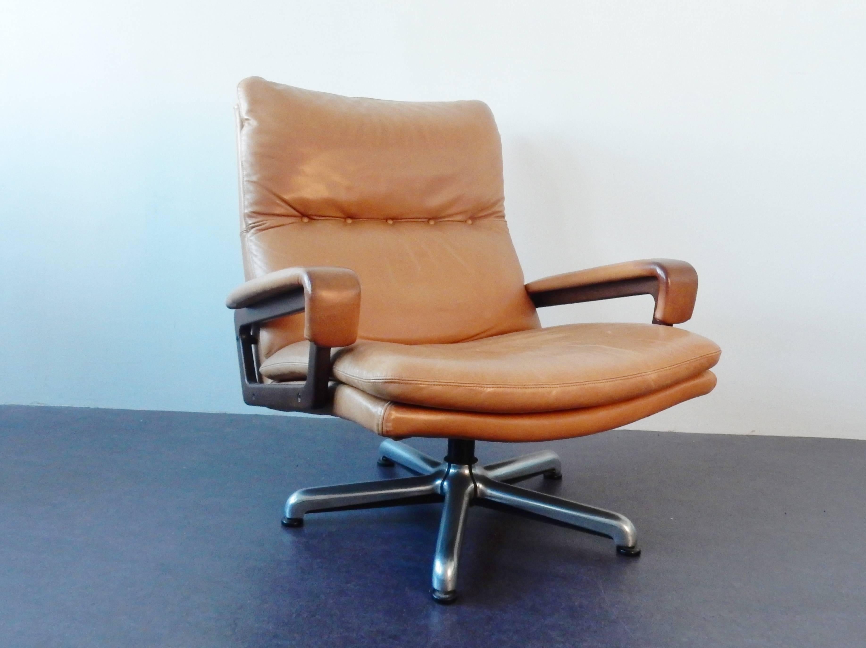 This cognac leather 'King' model lounge chair was made by Strässle and designed by André Vandenbeuck. The armchair has wooden armrests and stands on a metal swivel base. The leather does show some wear from age and use, but all is in a solid and