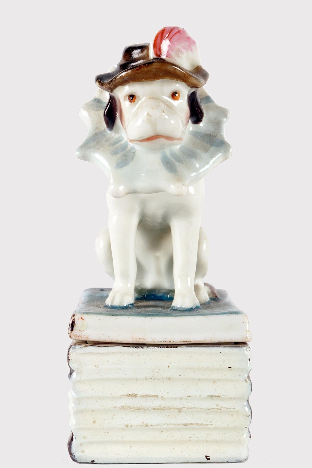 Rectangular box in painted porcelain, in the shape of stacked books. The base and lid are supported. Above the first book at the top, in the shape of a lid, there is a sitting dog, a King Charles Cavalier, with a plumed hat and ruff. The entire box
