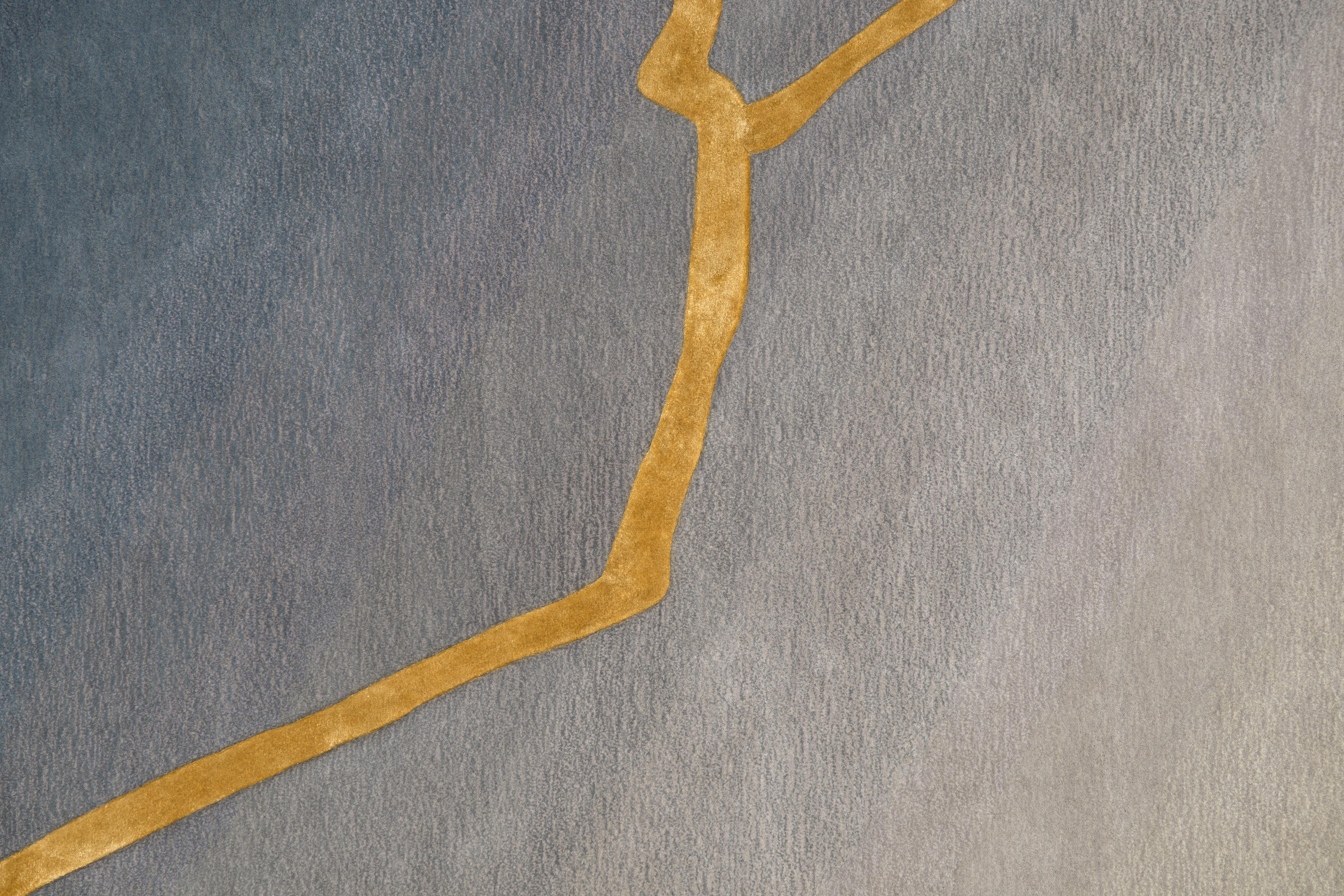 Der hier gezeigte handgetuftete Teppich K-3 weist einen raffinierten Verlauf von Grautönen auf, der nahtlos von tiefem Anthrazit zu einem weichen, silbernen Ton übergeht. Die Handwerkskunst wird durch ein verziertes goldenes, von Kintsugi
