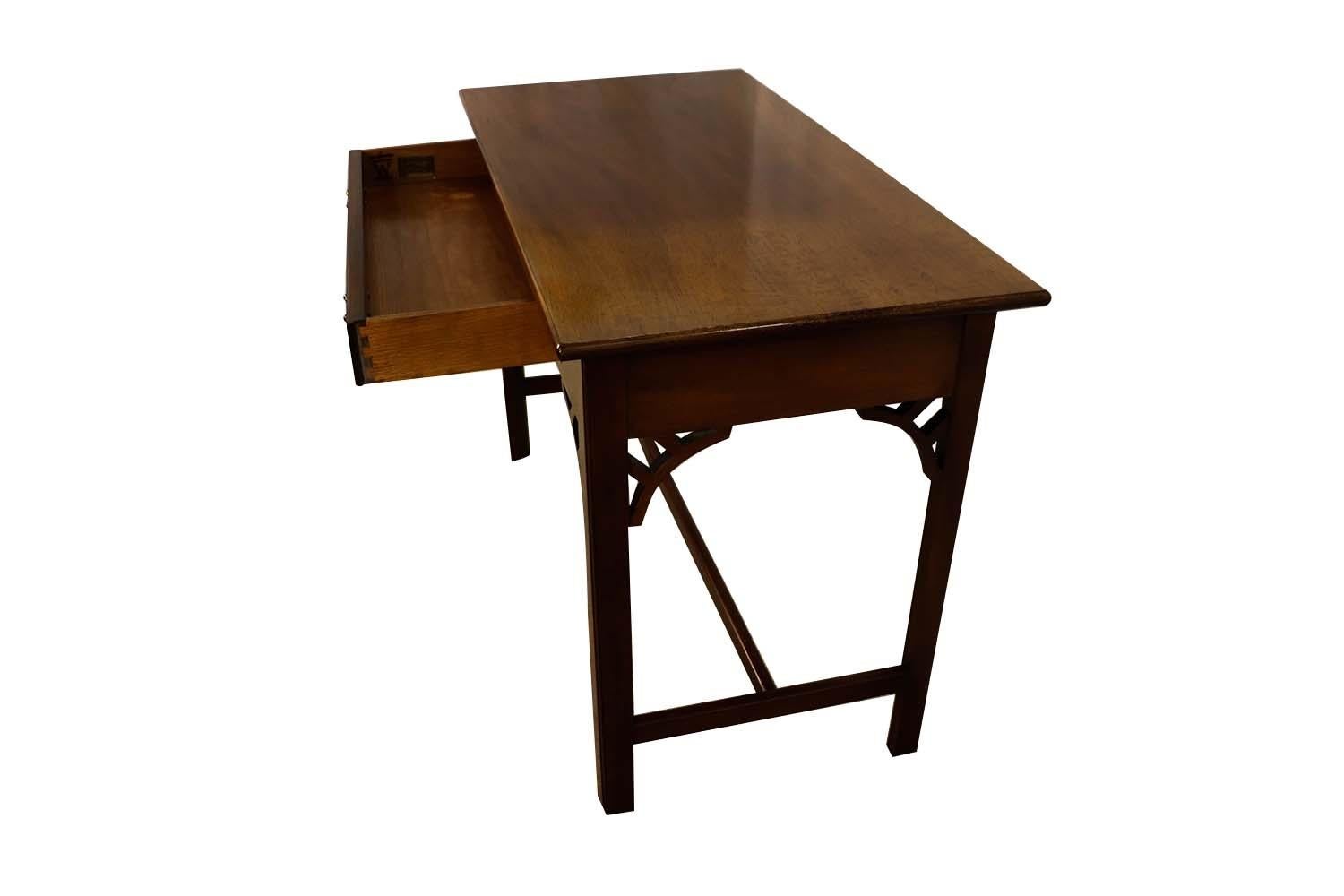 A solid mahogany hall console table or writing desk from the Williamsburg Adaptation collection by Kittinger Furniture Company of NY. This mahogany console table features a center drawer with four square legs with Chippendale fretwork attaching the