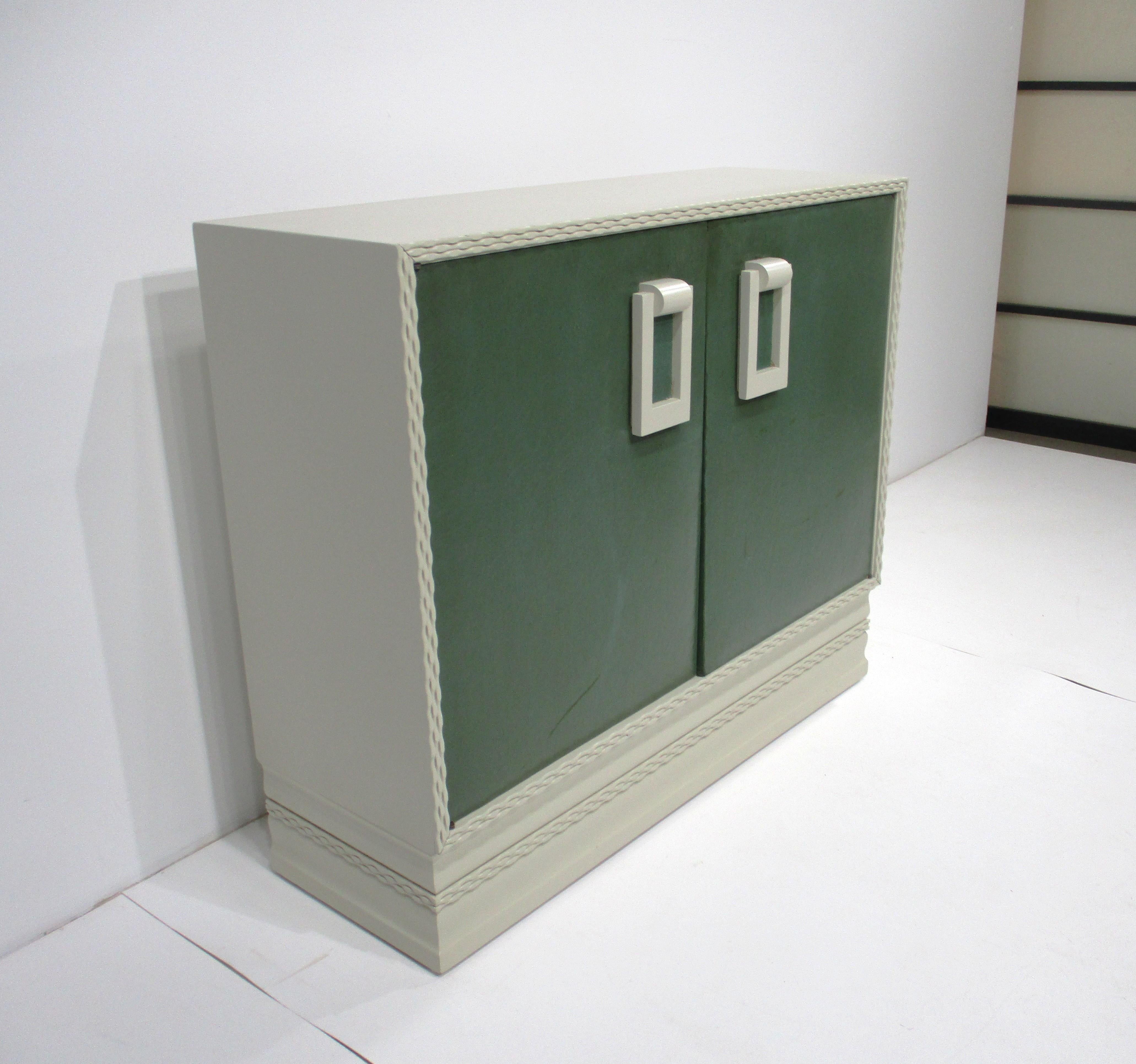 Una consolle/armadio a due ante in legno di colore bianco ecru che ricorda il lussuoso lino francese grezzo, con ante imbottite in tessuto ad olio di colore verde foglia. I tiranti in legno e gli eccezionali dettagli intagliati sulla parte anteriore