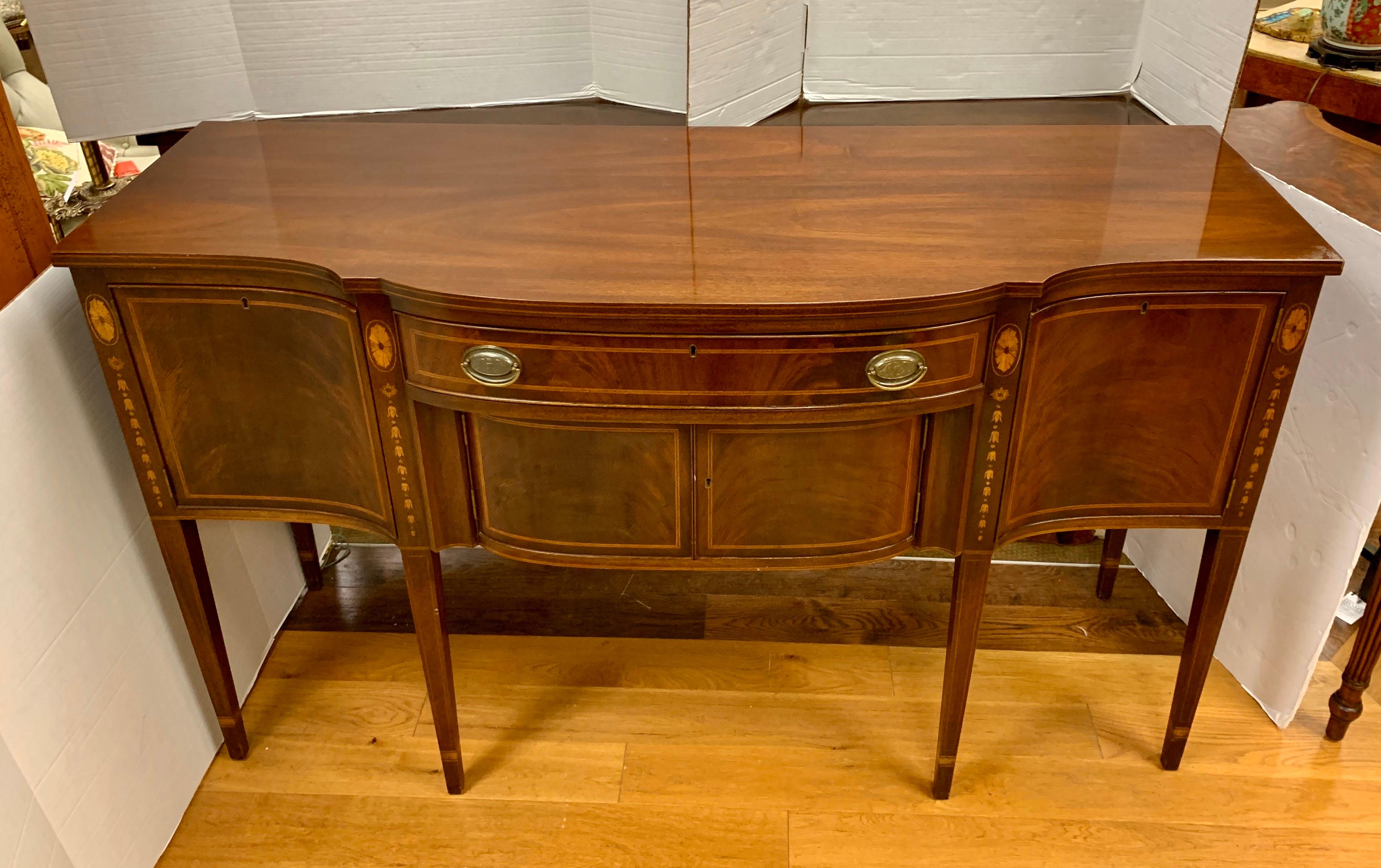 Outstanding large Sheraton bow front Kittinger sideboard constructed of solid mahogany and mahogany veneer. The matched flame mahogany veneer is highlighted with boxwood inlay and has beautiful bellflower inlay down the tapered legs. Brass American