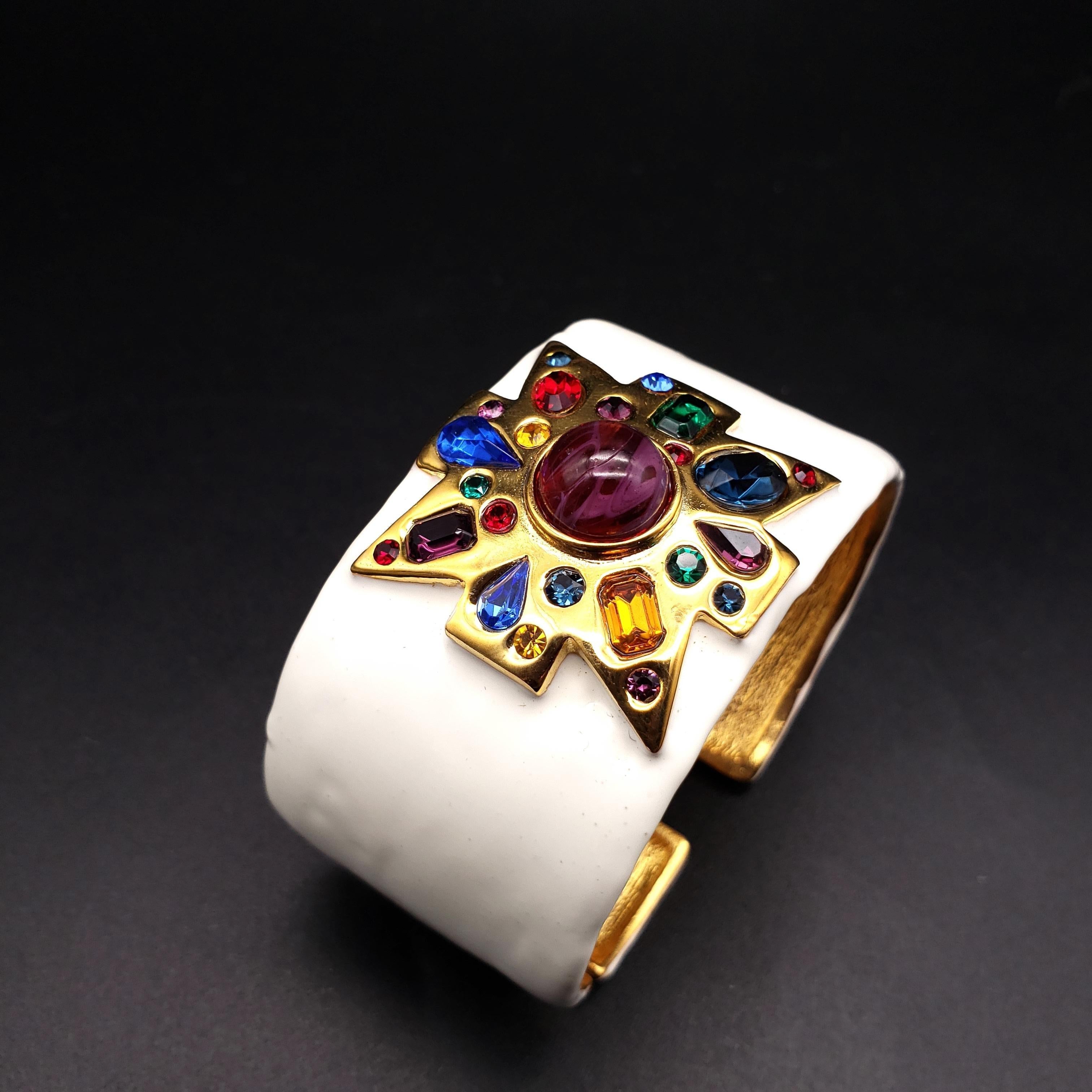 A bracelet by Kenneth Jay Lane. Painted white enamel, with a stylized maltese cross motif in gold. The centerpiece features a single teal-green resin cabochon, surrounded by clear crystals. Hinged bangle cuff.

Hallmarks: Kenneth © Lane
Inner