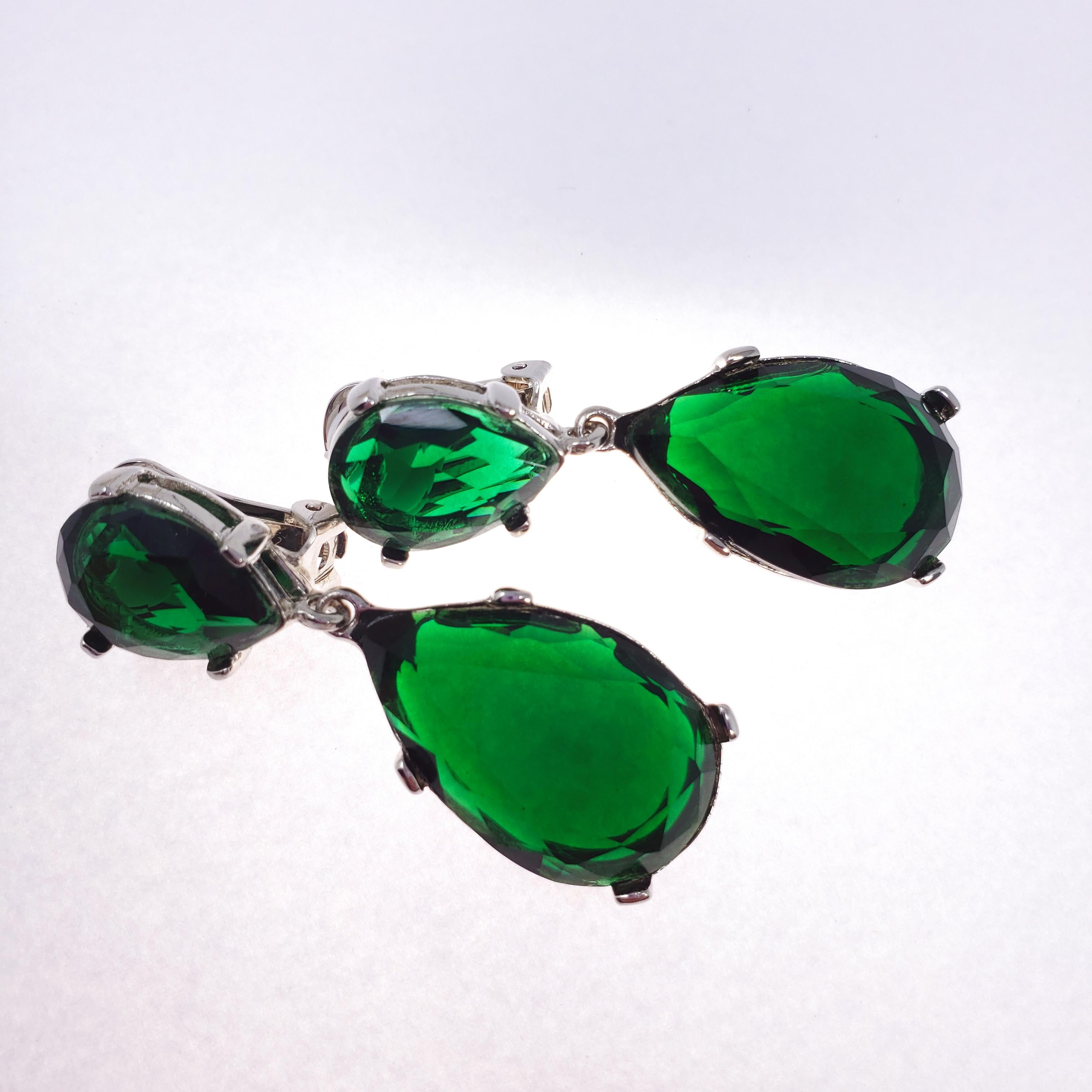 A pair of dangling clip on earrings by Kenneth Jay Lane. Each two teardrop-shaped emerald-colored crystals prong set in open back silver settings. Reflects an elegant, translucent green tone when under light, and a rich, dark green tone