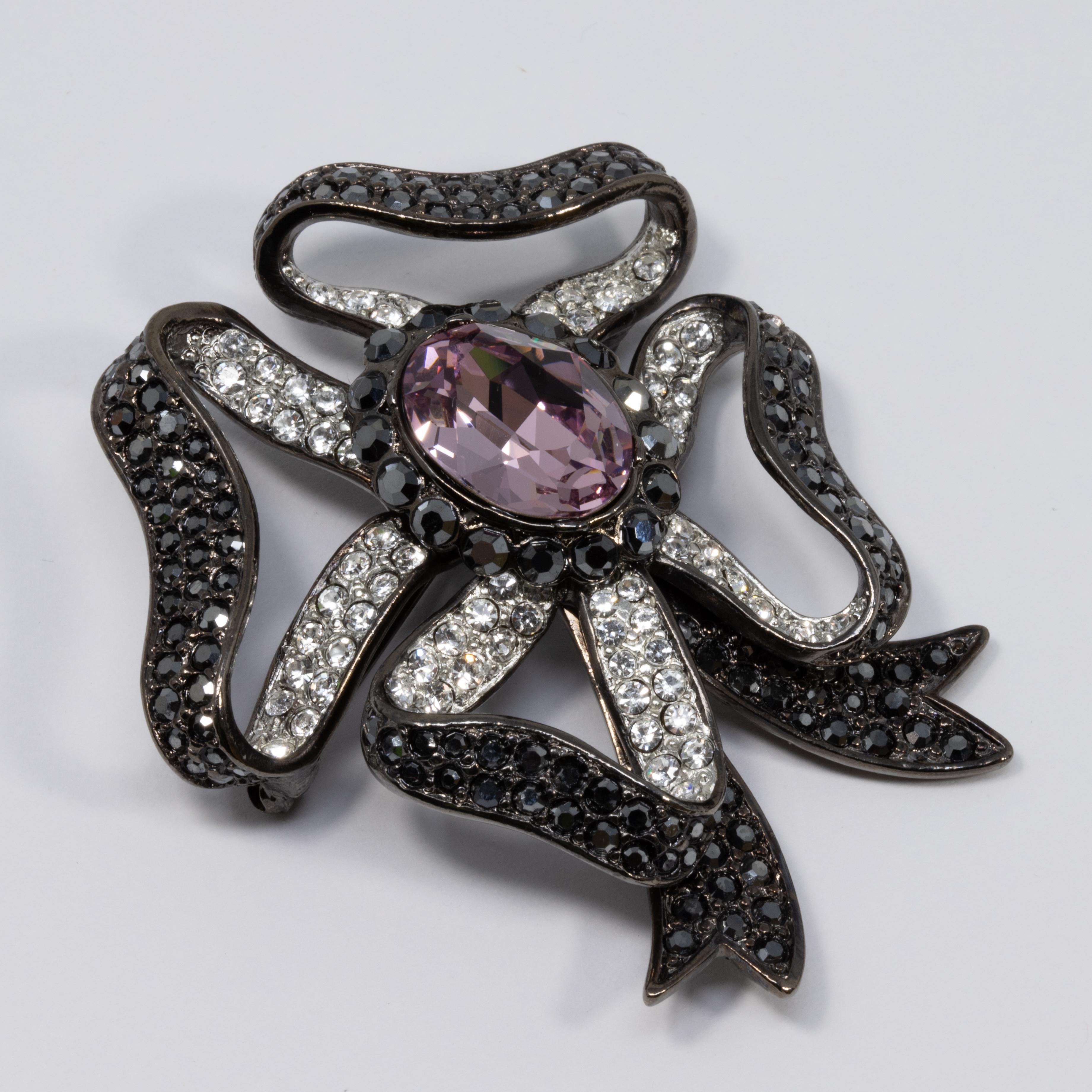 A stylish pin brooch by Kenneth Jay Lane! Features dark gunmetal tone bow accented with clear and dark iridescent pave crystals. A bright amethyst crystal is set in the center for a bold contrast.

Hallmarks: Kenneth © Lane