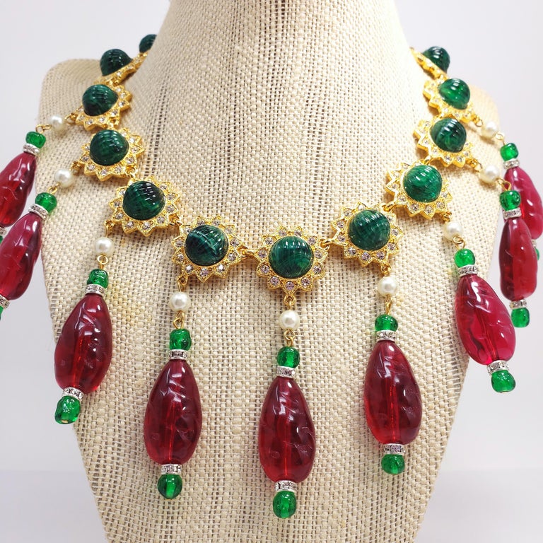 An ornate necklace from Kenneth Jay Lane. Features emerald green textured resin accents, set in gold plated floral setting and accented with clear crystals. Carved ruby red resin accents hang off the necklace, accented with pearls and silvertone