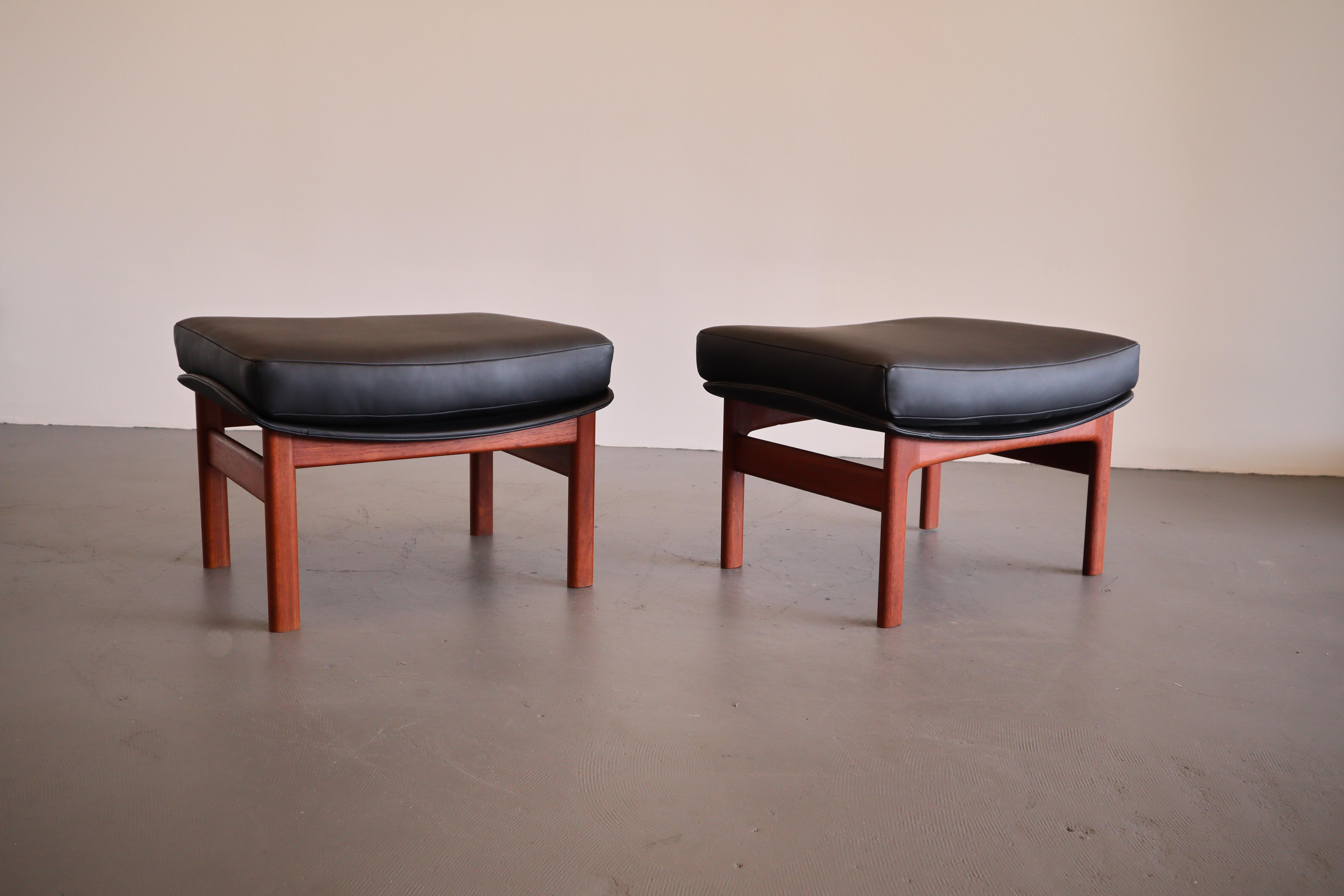 Minimalist and organic, Larsen's design ethos incorporated functionality and high quality materials. An exceptional pair of midnight black leather ottomans for Mogens Kold Møbelfabrick. The upholstery compliments the robust teak base and pleasant