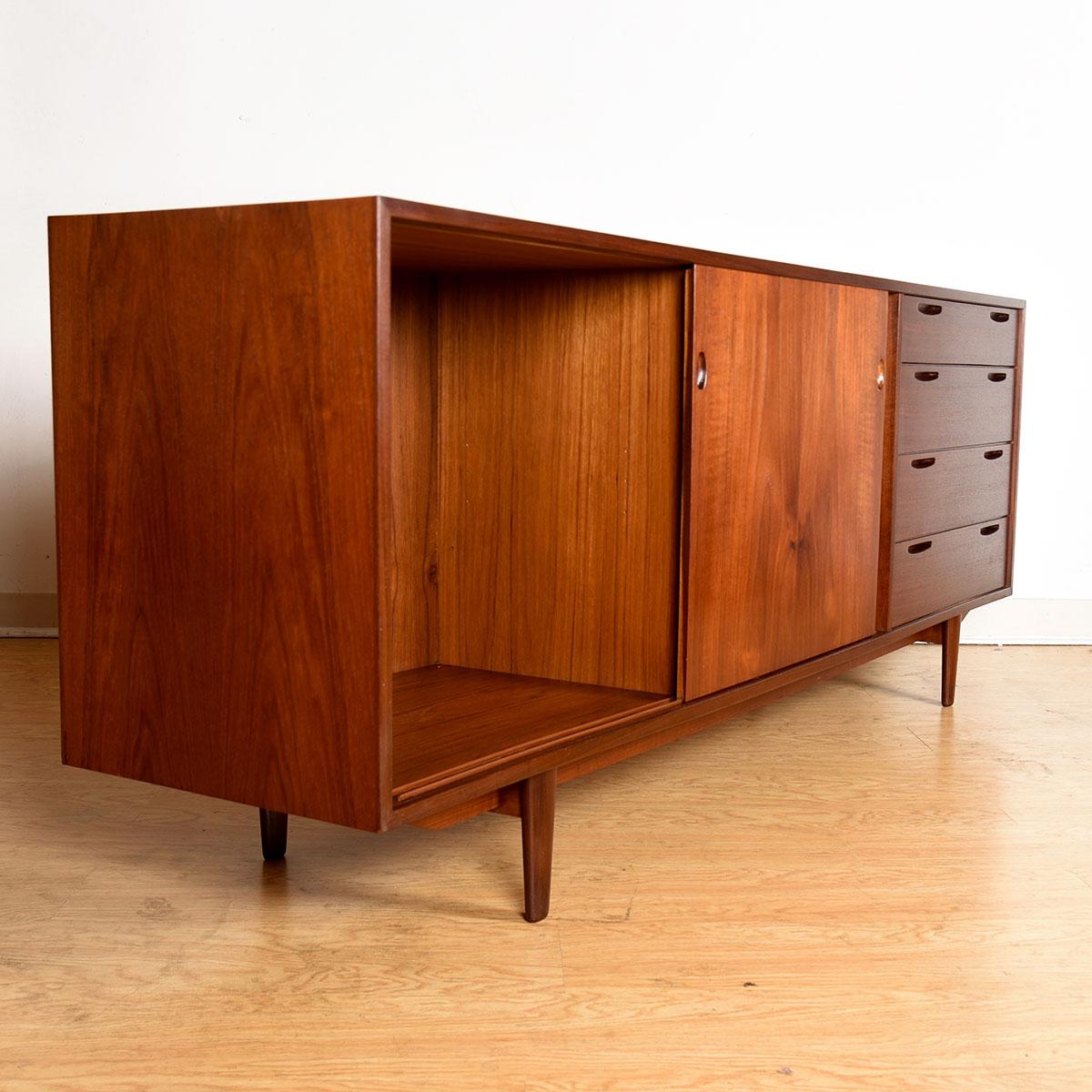 Stunning Danish modern credenza in deep dark teak by Kofod-Larsen.
Column of drawers on the far right, each having two narrow slits framed in darker teak for pulls.

Two sliding doors open to cabinet storage with an open bay on the left.
Stretcher