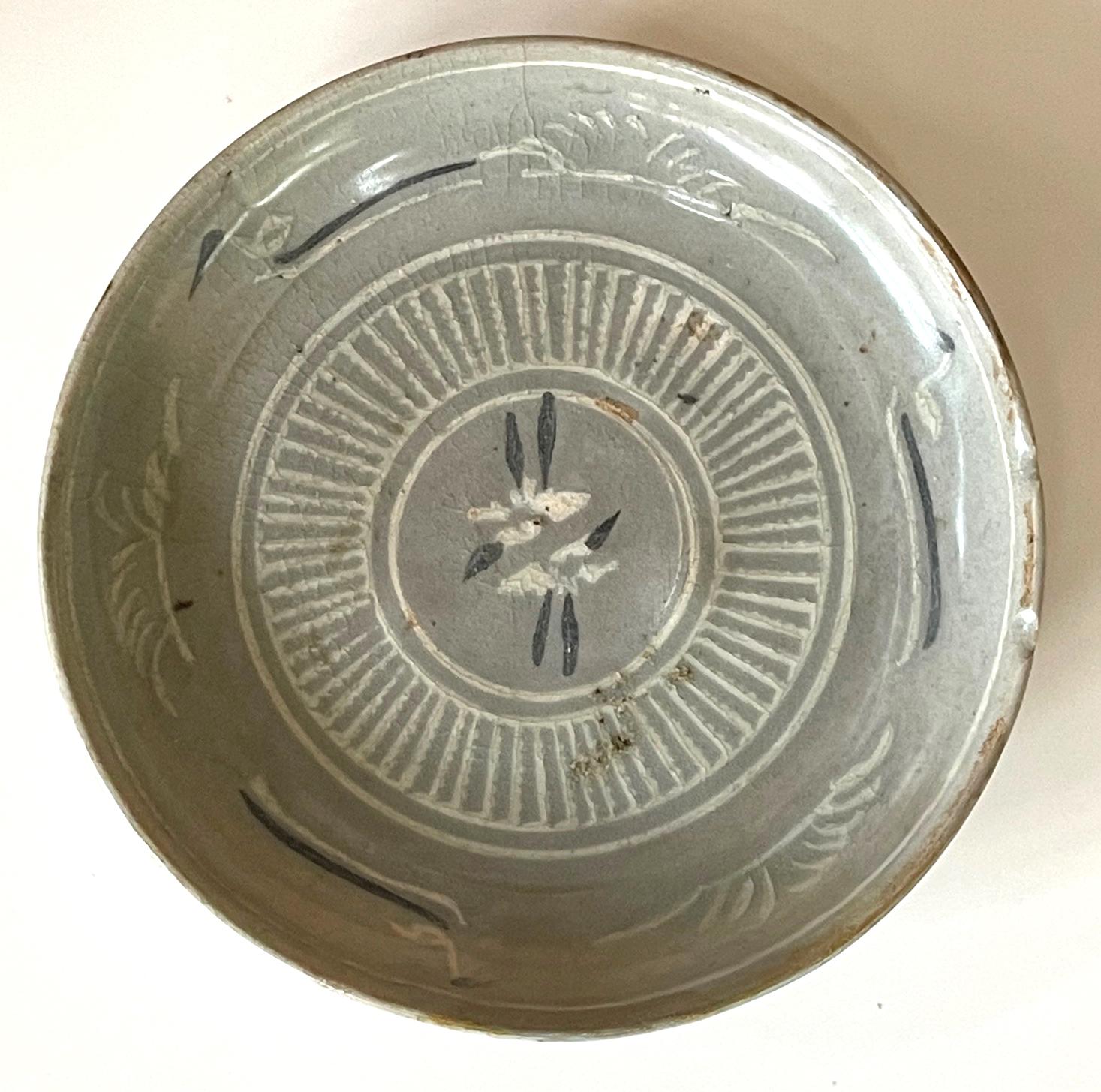 The round ceramic plate with a slightly raised foot ring is dated from late Korean Goryeo Kingdom (918 to 1392 AD) likely toward the end of the 14th century. The plate features a celadon glaze and underglaze slip inlays in black and white. The