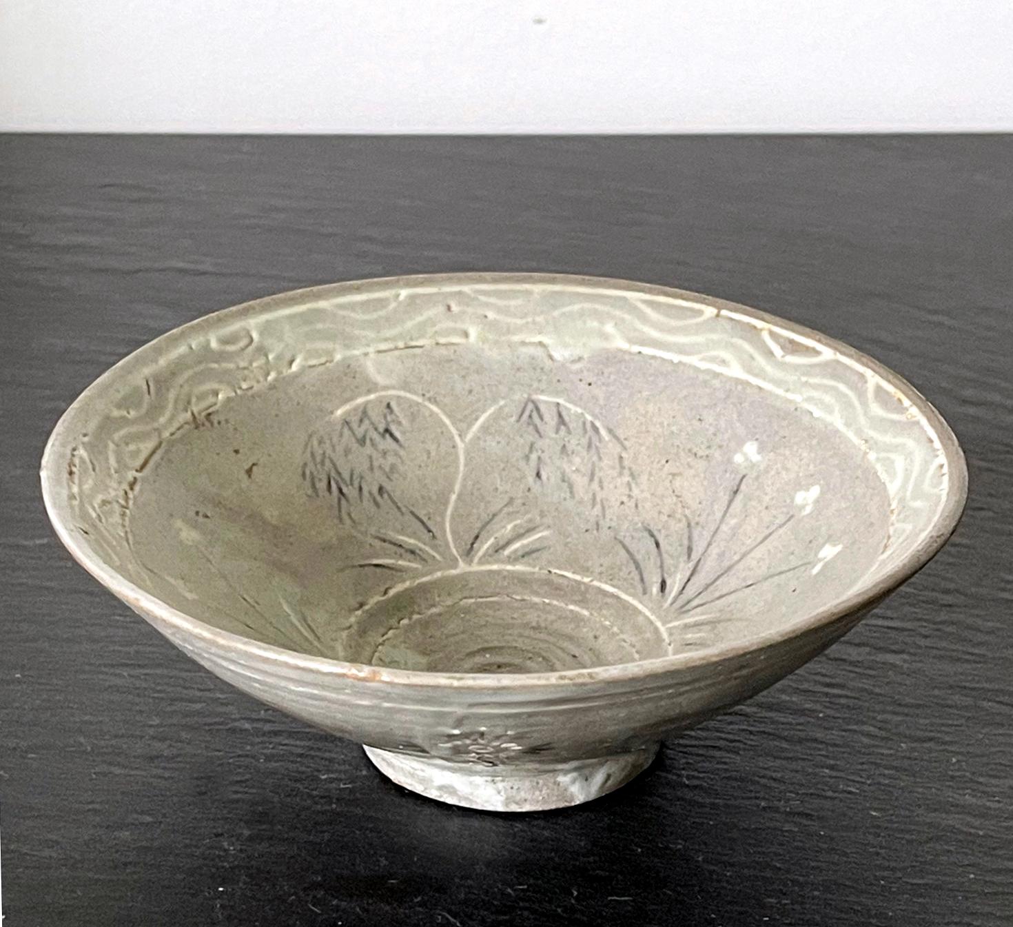 A small ceramic conical form tea bowl from Korean Goryeo Dynasty circa 14th century. The bowl is of a slightly irregular shape and covered in a grayish overglaze. The inlaid slip decoration in black and white features willow trees with weeping