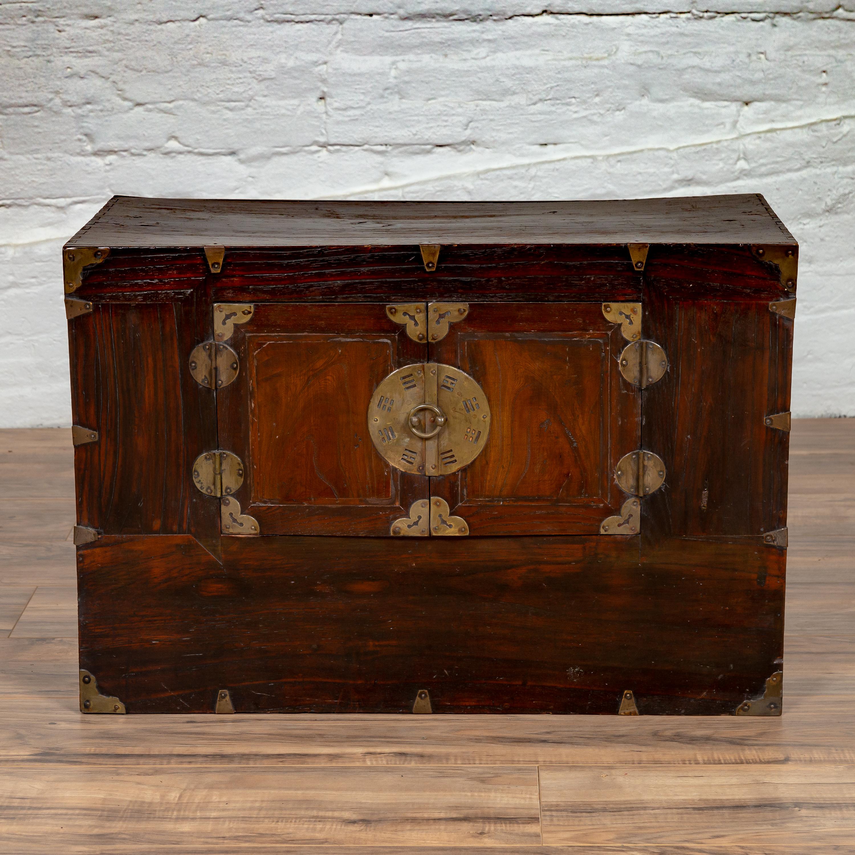 A Korean elmwood two section clothing cabinet from the late 19th, early 20th century, with double doors, antique brass accents and round medallion lock. This lovely Korean cabinet features a rectangular top sitting above a two section façade, made