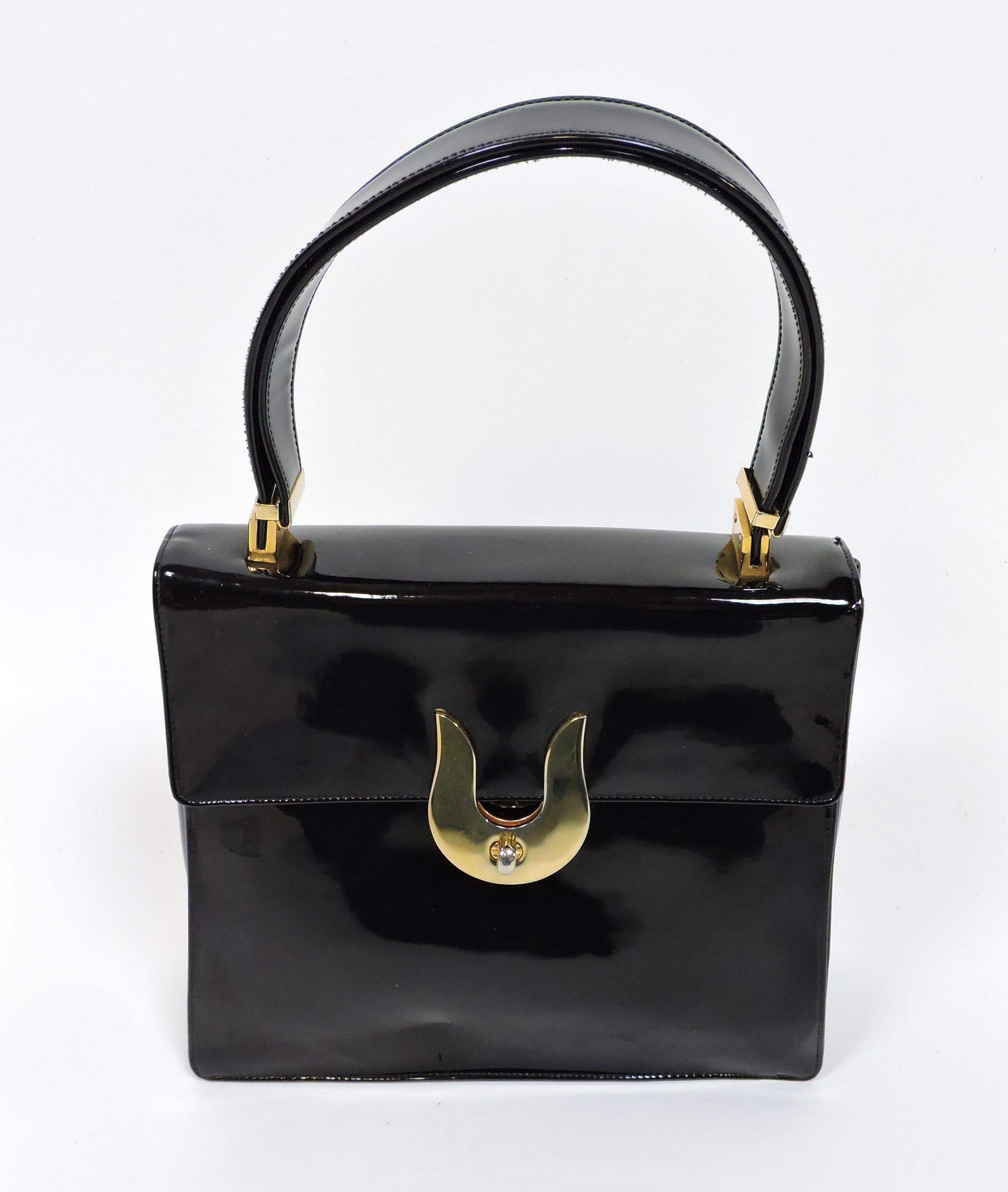 Stylish black patent leather adjustable handbag made by high end manufacturer, Koret. This bag has a fold over flap with an unusual brass U-shaped twist clasp closure. The interior is lined with leather and has a pocket with a chain purse tethered