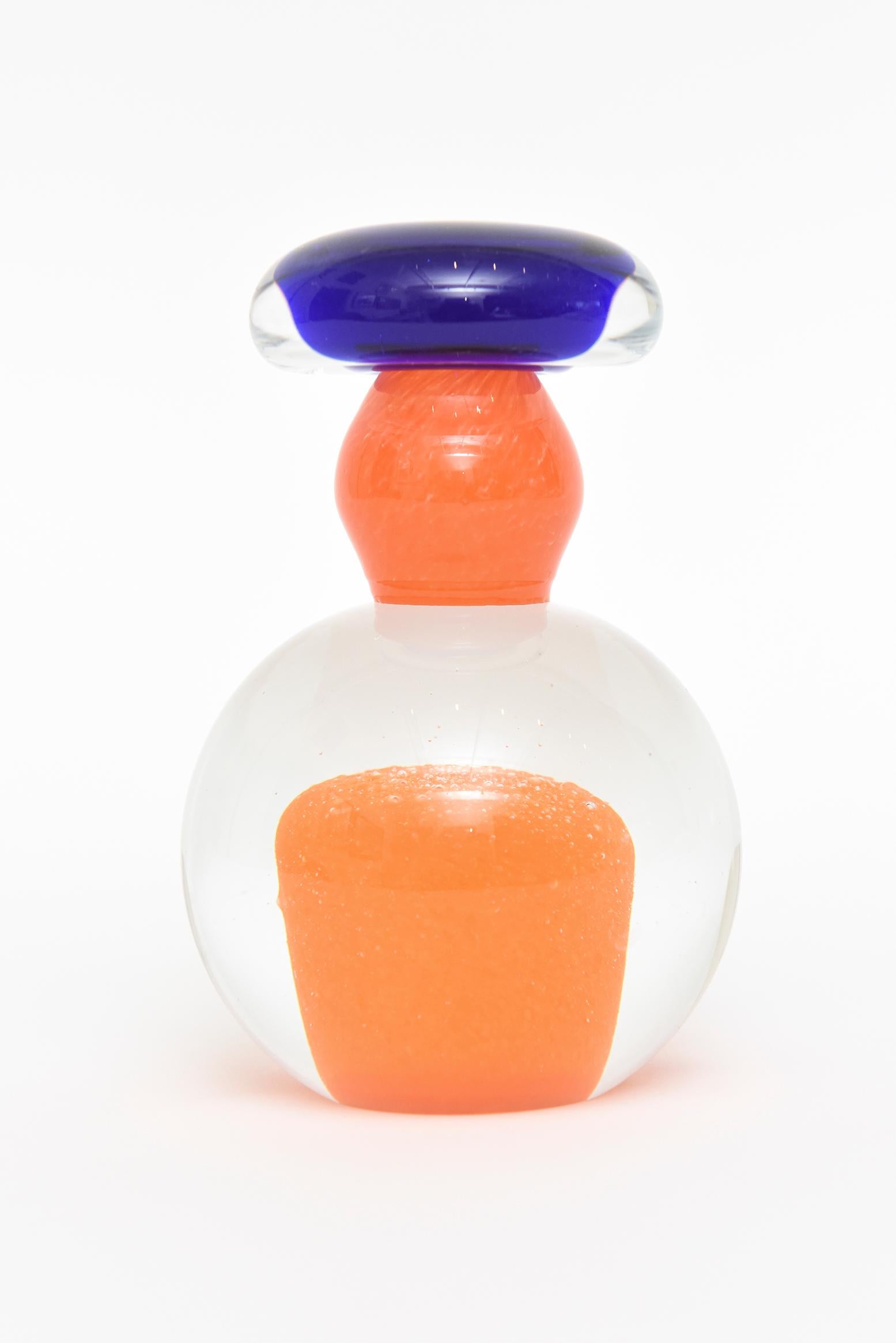This Kosta Boda glass paperweight sculpture has immersions of an orange blob floating inside the clear. It has tiny dots on top and looks like an orange marshmallow. The mottled orange neck is typical of Kosta Boda. The cobalt blue top pops! The