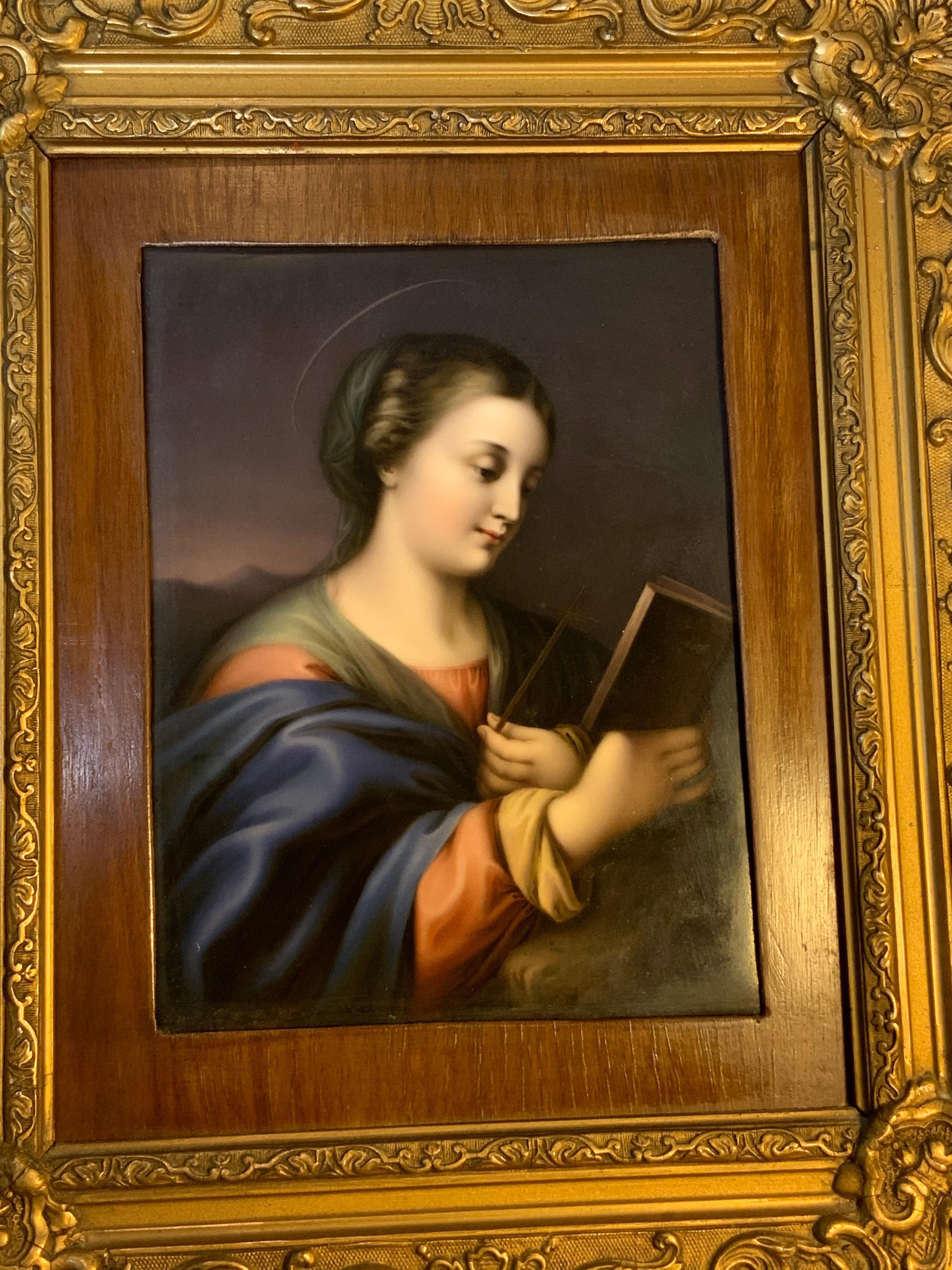 This is a KPM style hand painted plaque of Saint Catherine of Alexandria. The bright colors of her robes highlight her porcelain skin. She appears holding a book with her right hand & a pen in the other hand. The plaque has a carved plaster and wood