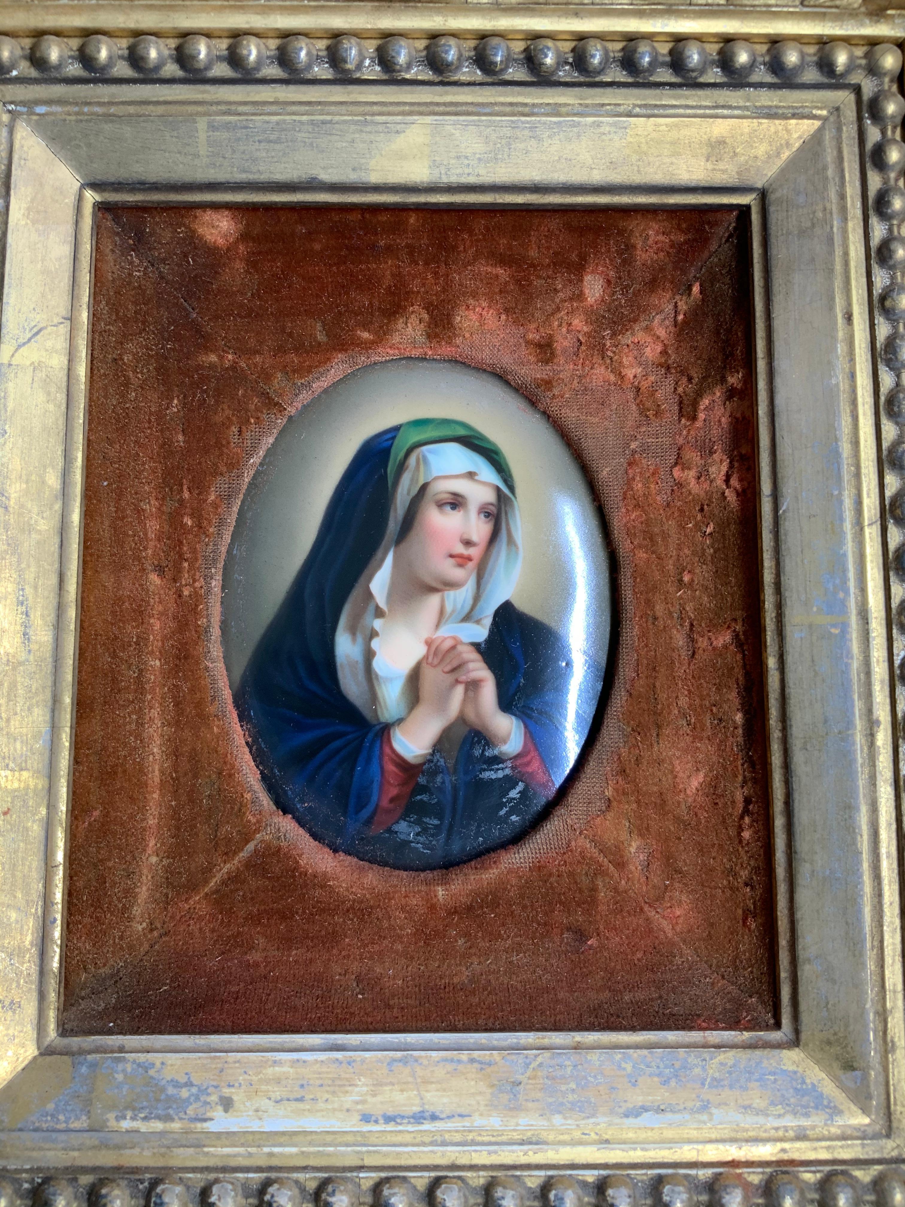 This is a hand painted porcelain oval plaque portrait of the Virgin Mary praying. It is mounted in a carved gilded wood frame embellished with gilt scrolls of acanthus, bay leaves bunches and a chain of beads.