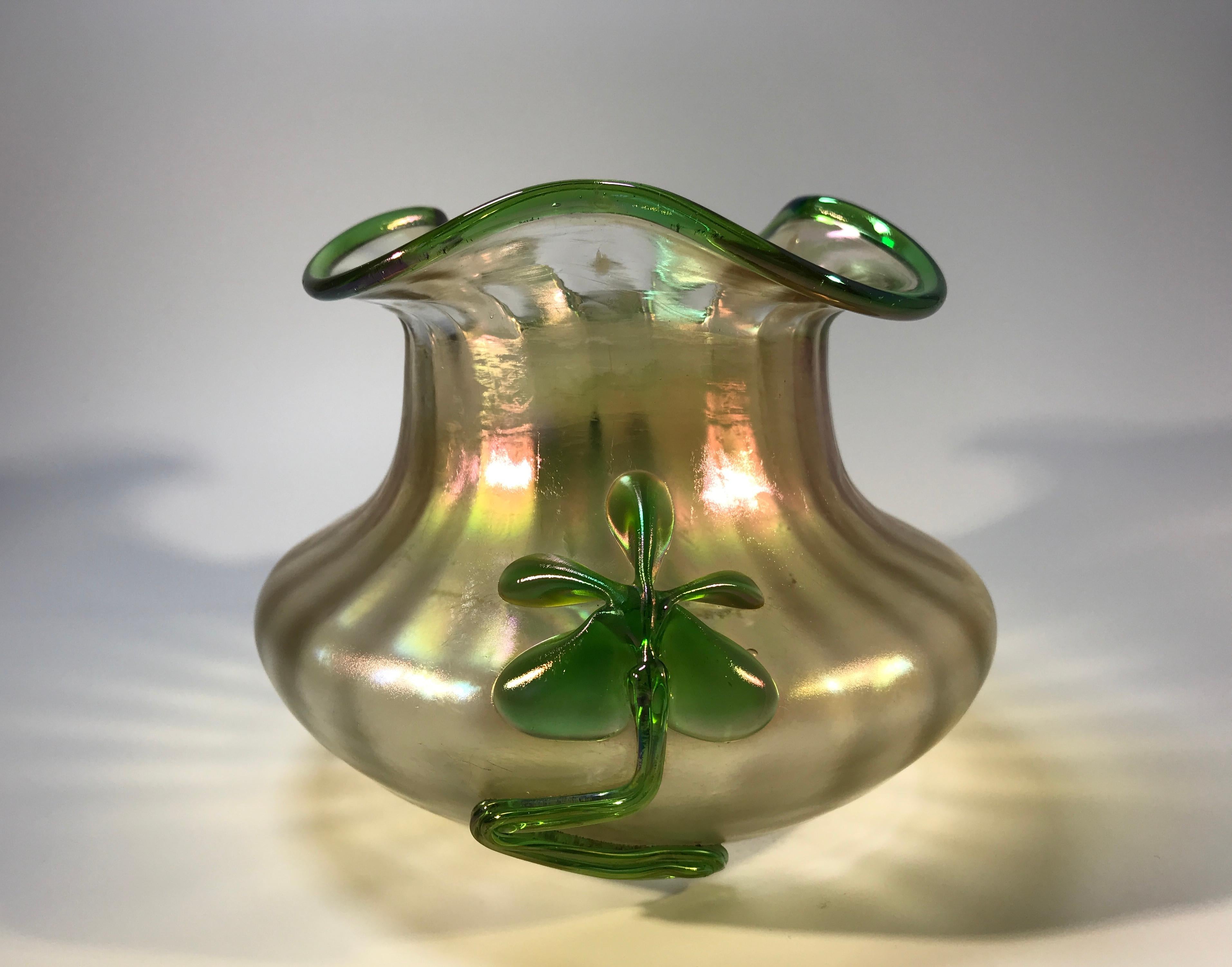 Vase en verre organique irisé, soufflé à la main, de style Art déco. Décoration appliquée de deux motifs floraux verts de part et d'autre et bordure verte sur le bord. La base présente un pontil poli à la meule, preuve de l'art des souffleurs de