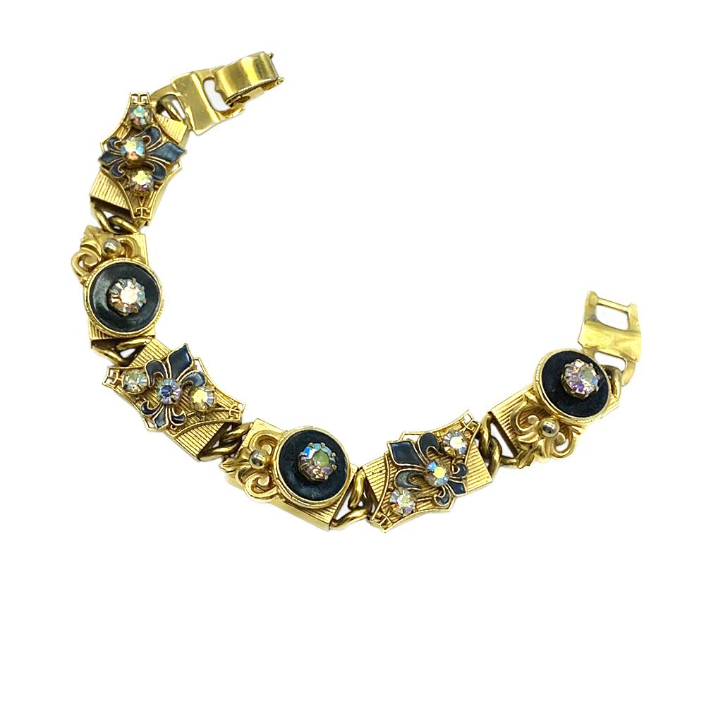 This is a 1960s Kramer of NY bracelet with Fleur de Lis. It has six linked detailed 3D design gold tone metal block sections and a fold-over clasp. Alternating sections are decorated with fleur di lis and round black enamel button shape with prong