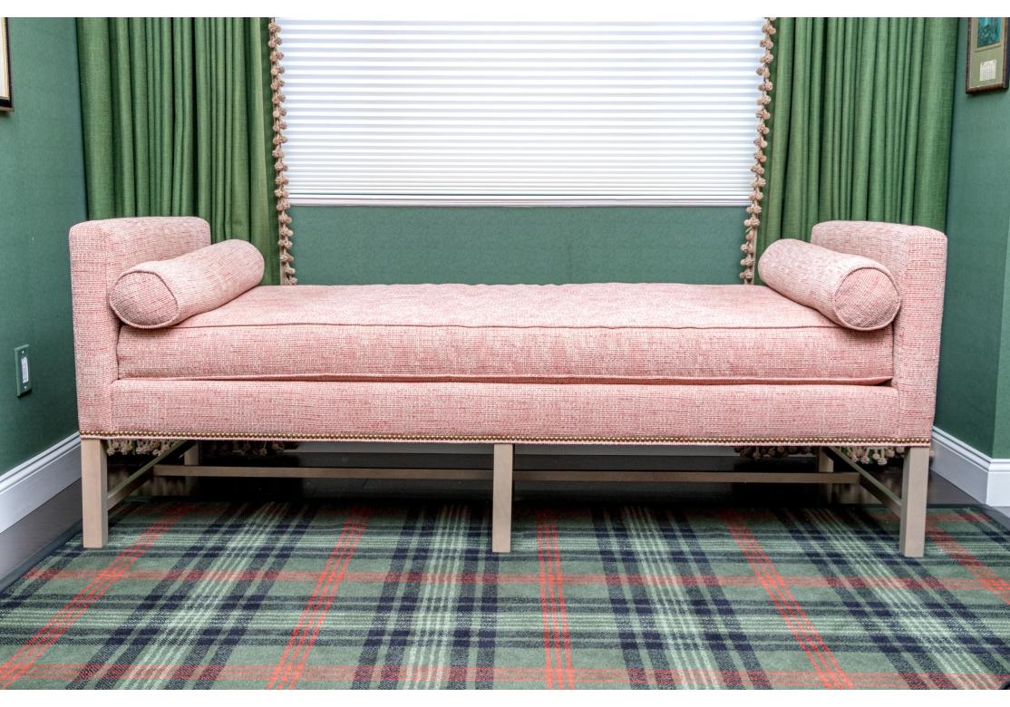 An upholstered day chaise lounge by Kristin Drohan with clean lines, wood frame with double H form stretcher, straight arms, bolster pillows at both ends. Upholstered in a red and tan textured fabric and contrasting tan zippers. The seat and pillows
