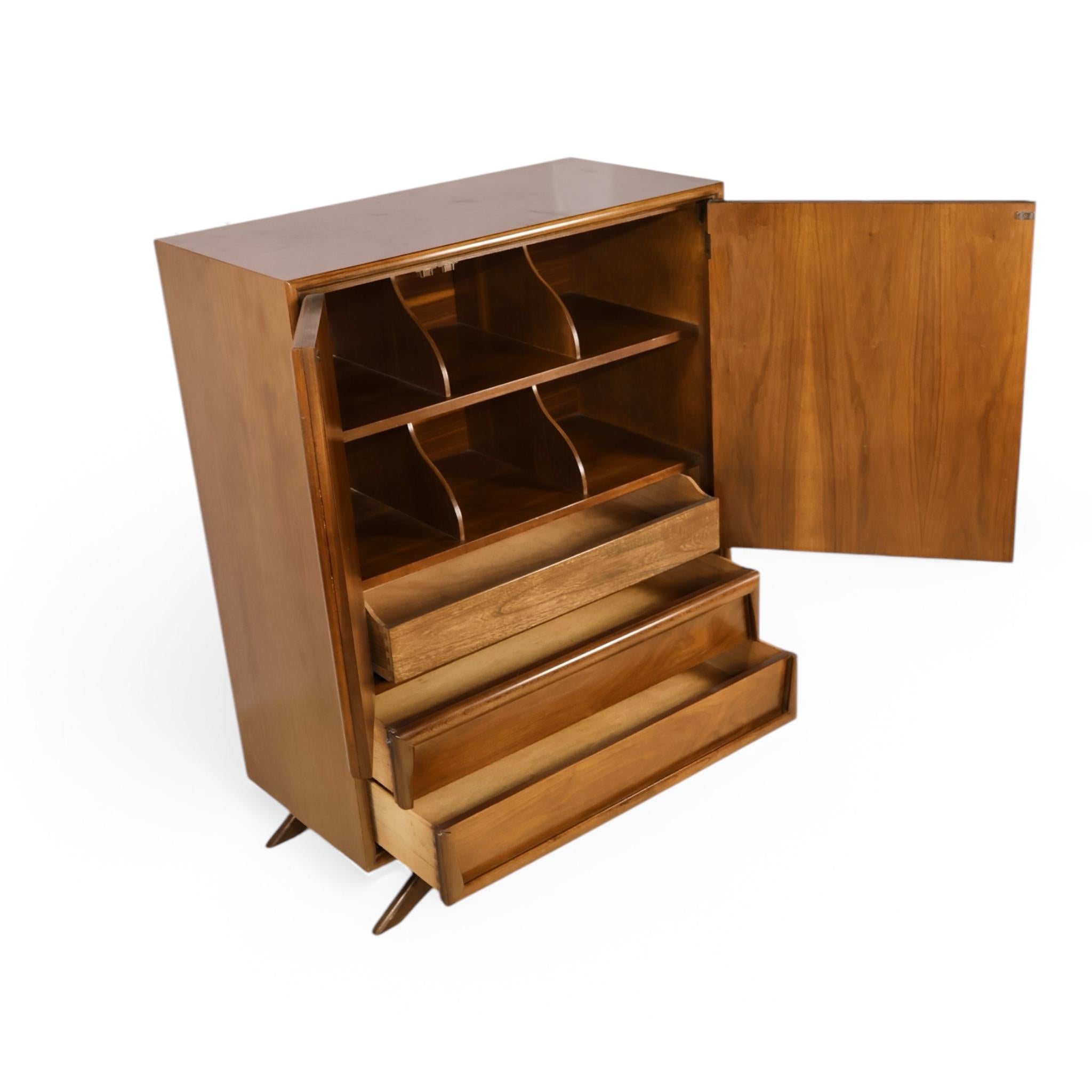 This vintage mid-century modern highboy dresser by Kroehler showcases bold sculptural lines and striking geometric detailing that capture the essence of the era’s design. The front features three carved panel doors with rich walnut grain, opening to