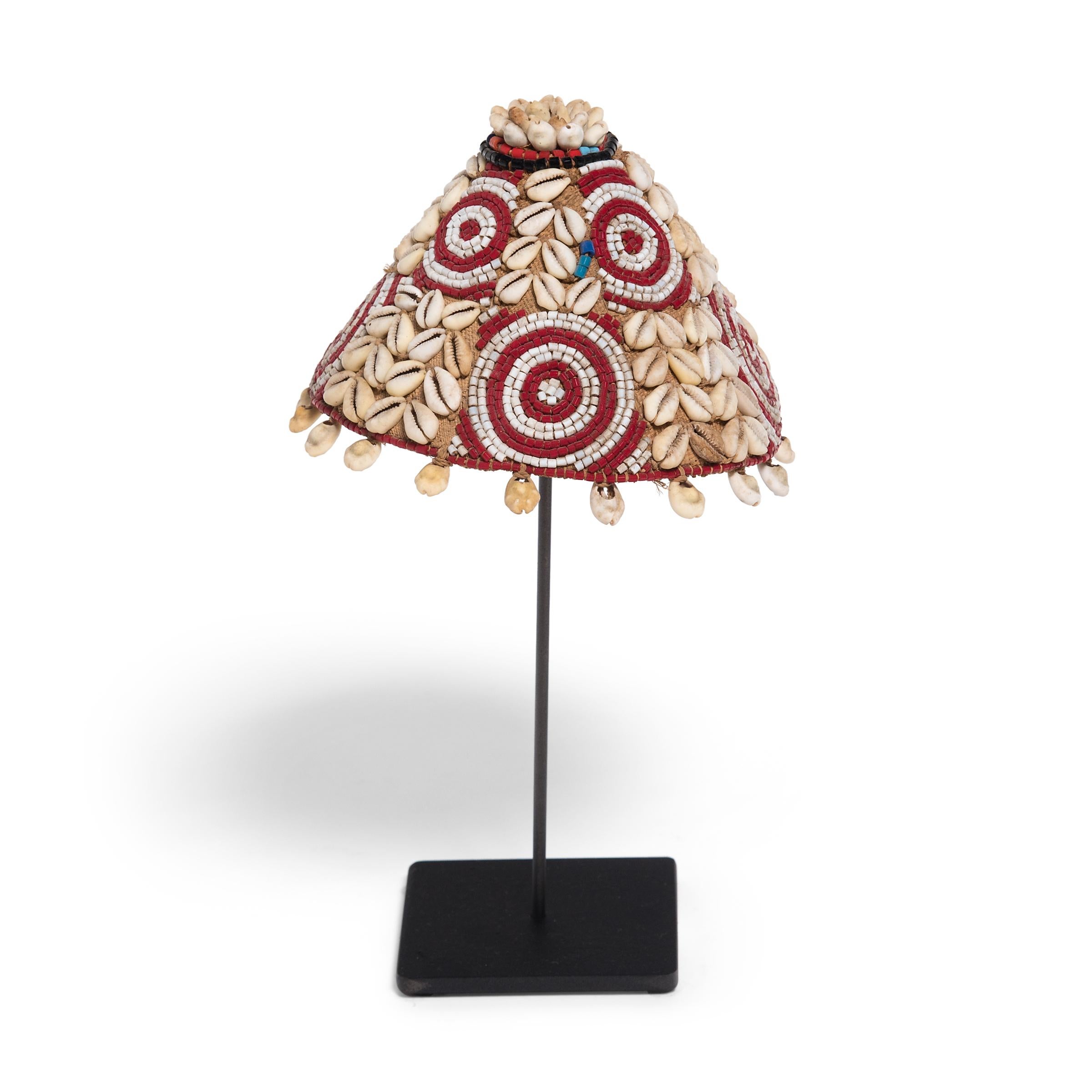 This petite cone-shaped cap, known as laket, was created by an artisan of the Kuba peoples of the Democratic Republic of the Congo. The base is made of a simple woven raffia fabric, which was then embellished with a pattern of cowrie shells and